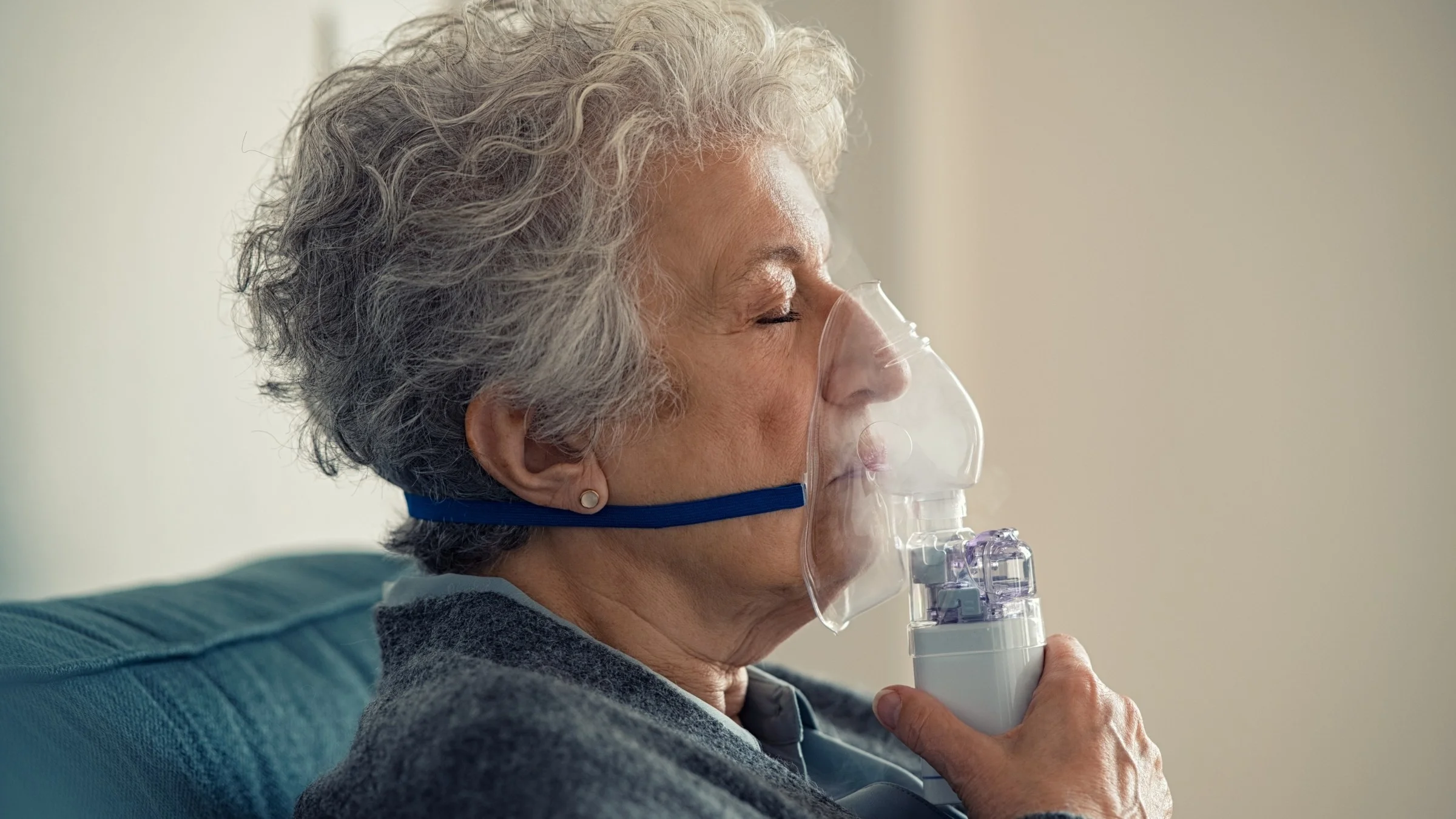 Close-up of an elderly woman using an over the nose nebulizer at home on the couch.
