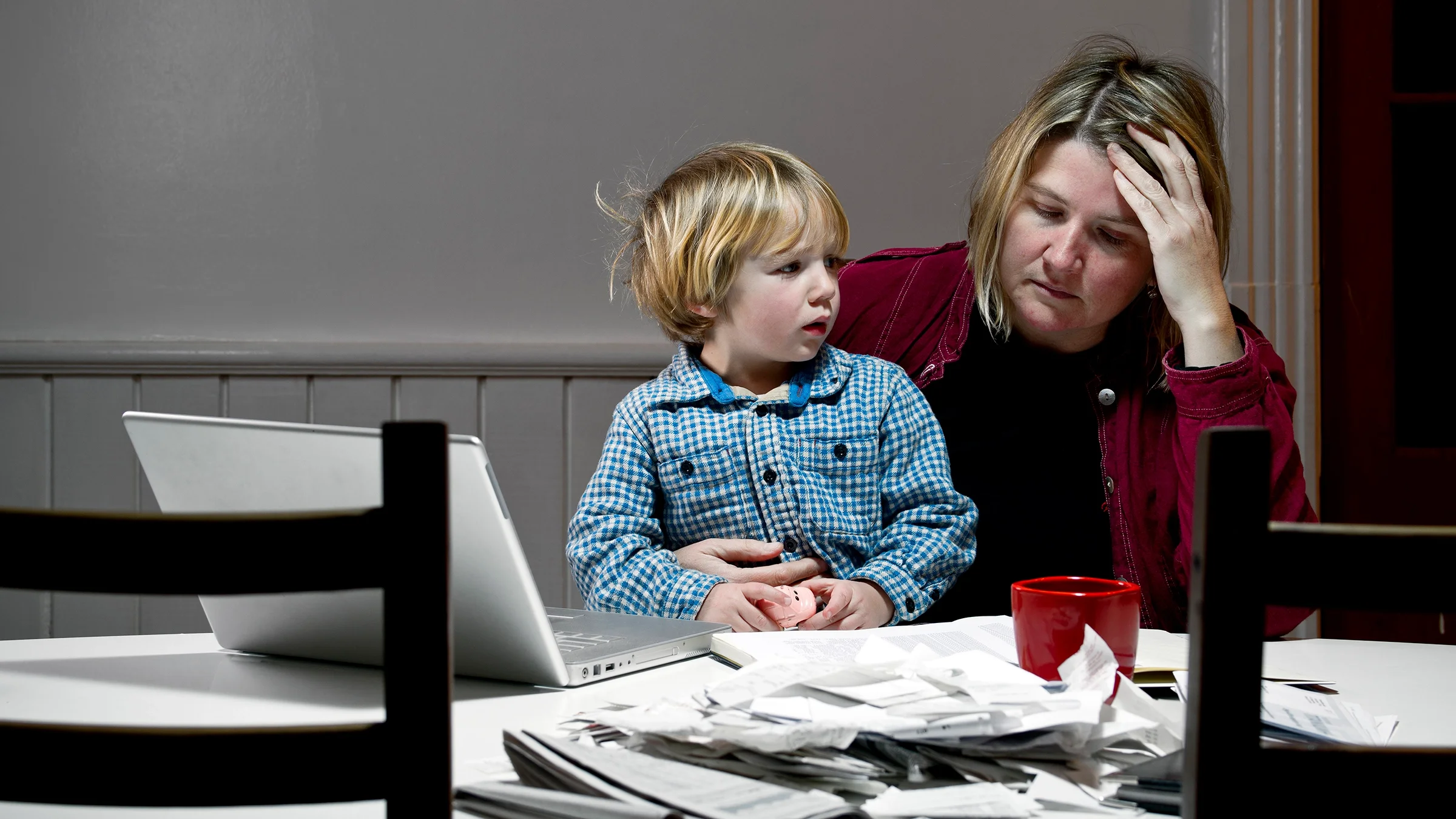 Mother holding her child in her lap as she reviews paperwork and finances. She has her hand on her forehead and looks frustrated and sad.