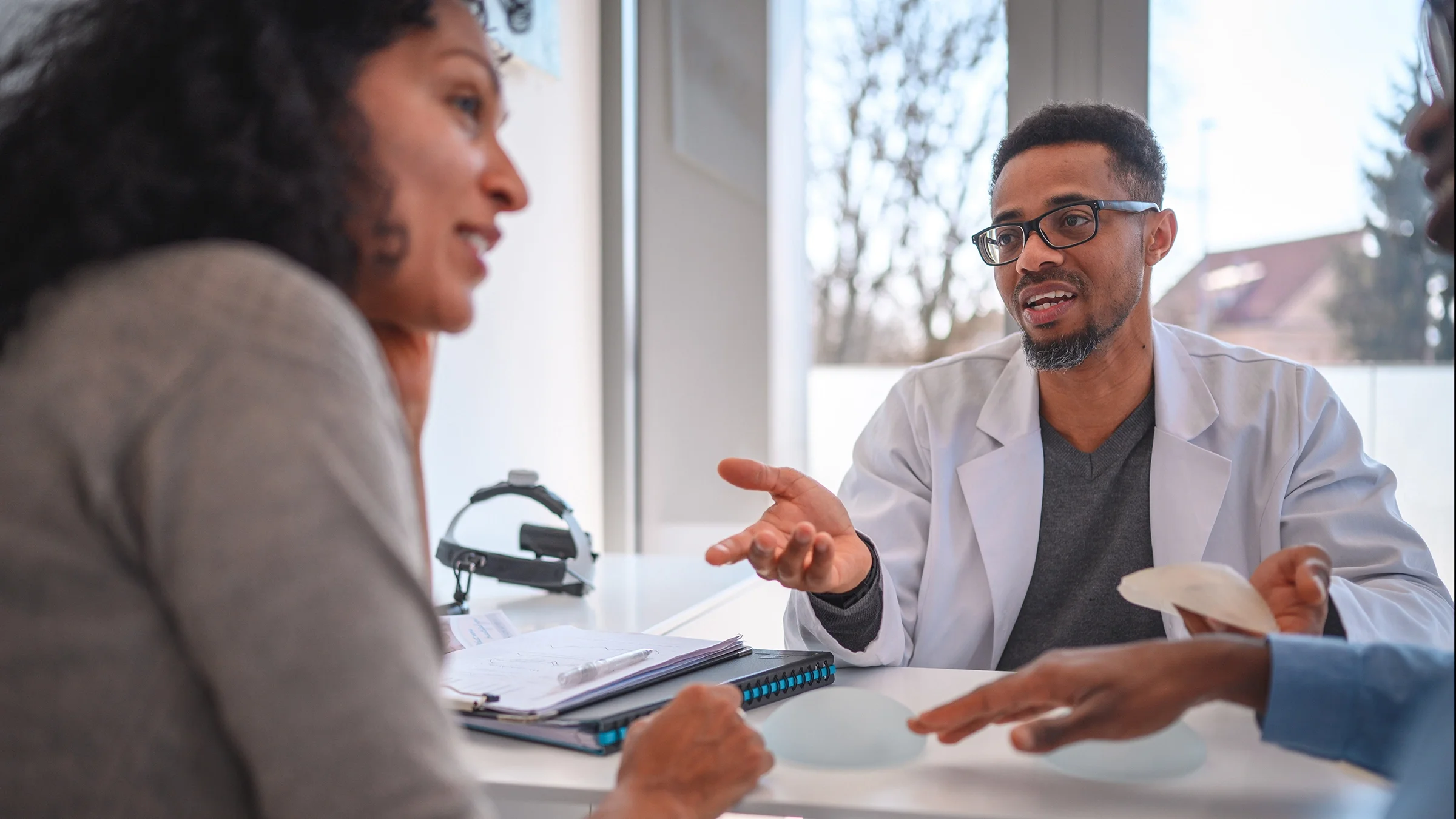 Young woman having a breast augmentation consultation with her doctor. There is a silicon breast implant on the desk.