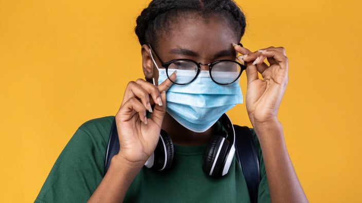 Young girl with itchy watery eyes on a yellow background. She has headphones around her neck and a face mask on.