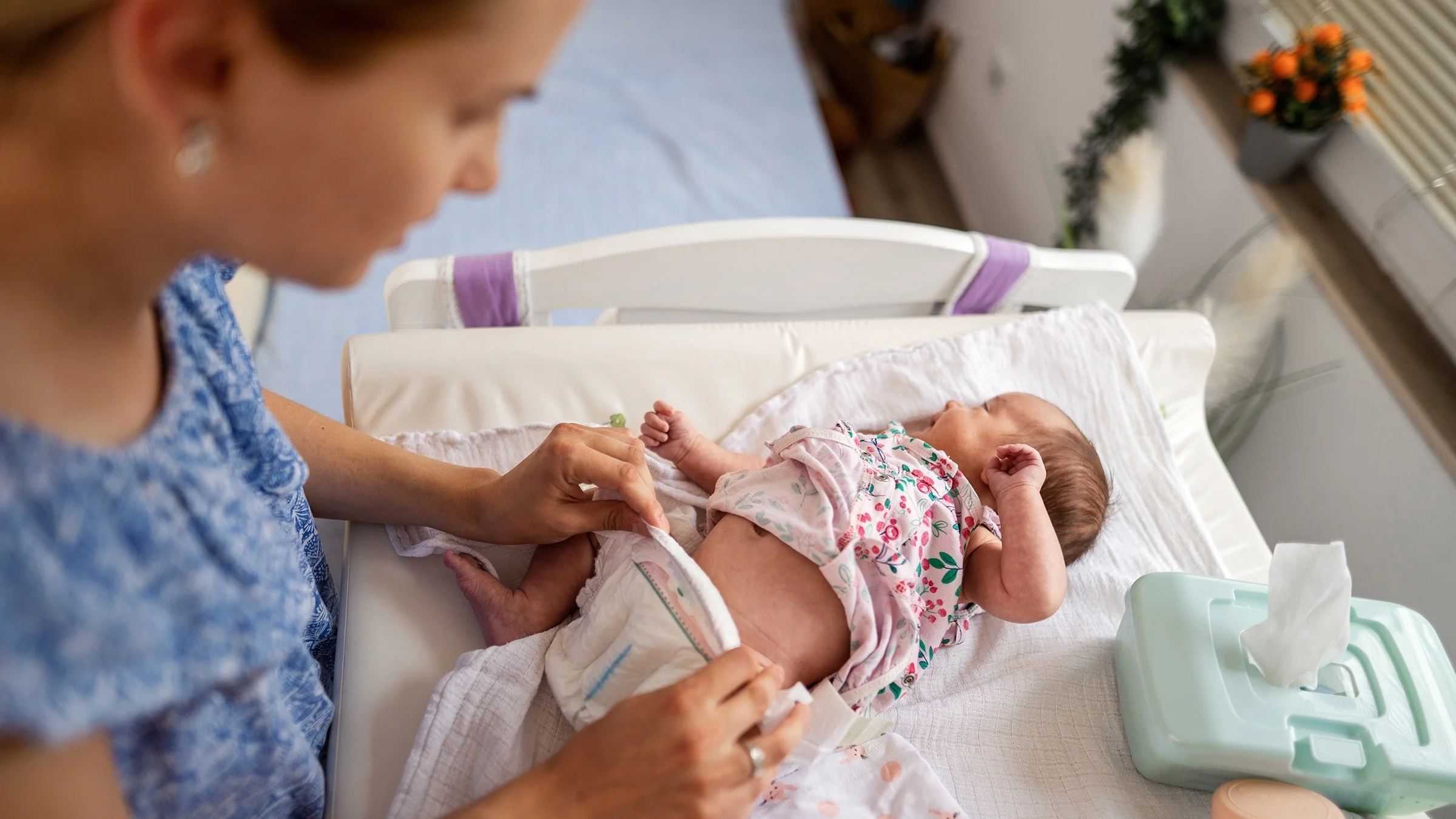 Mother changing a newborn's diaper on a changing table. The angle is shot from above with the focus on the baby. The mother is out of focus and blurry.