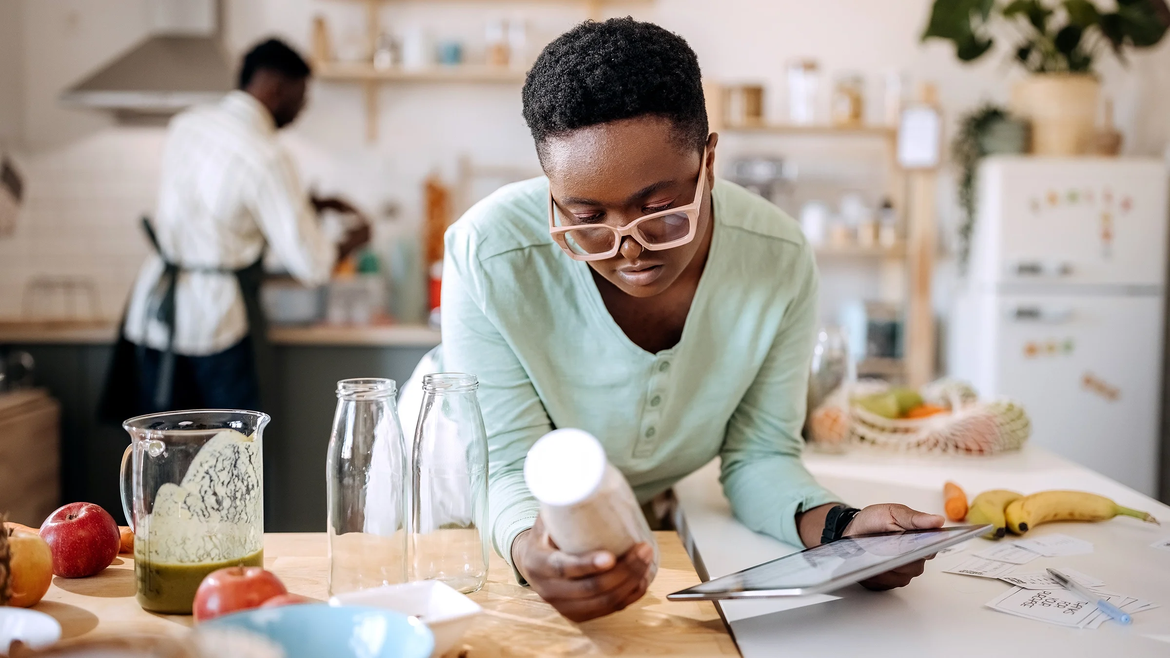 Woman reading a tablet and making a smoothie in a kitchen.