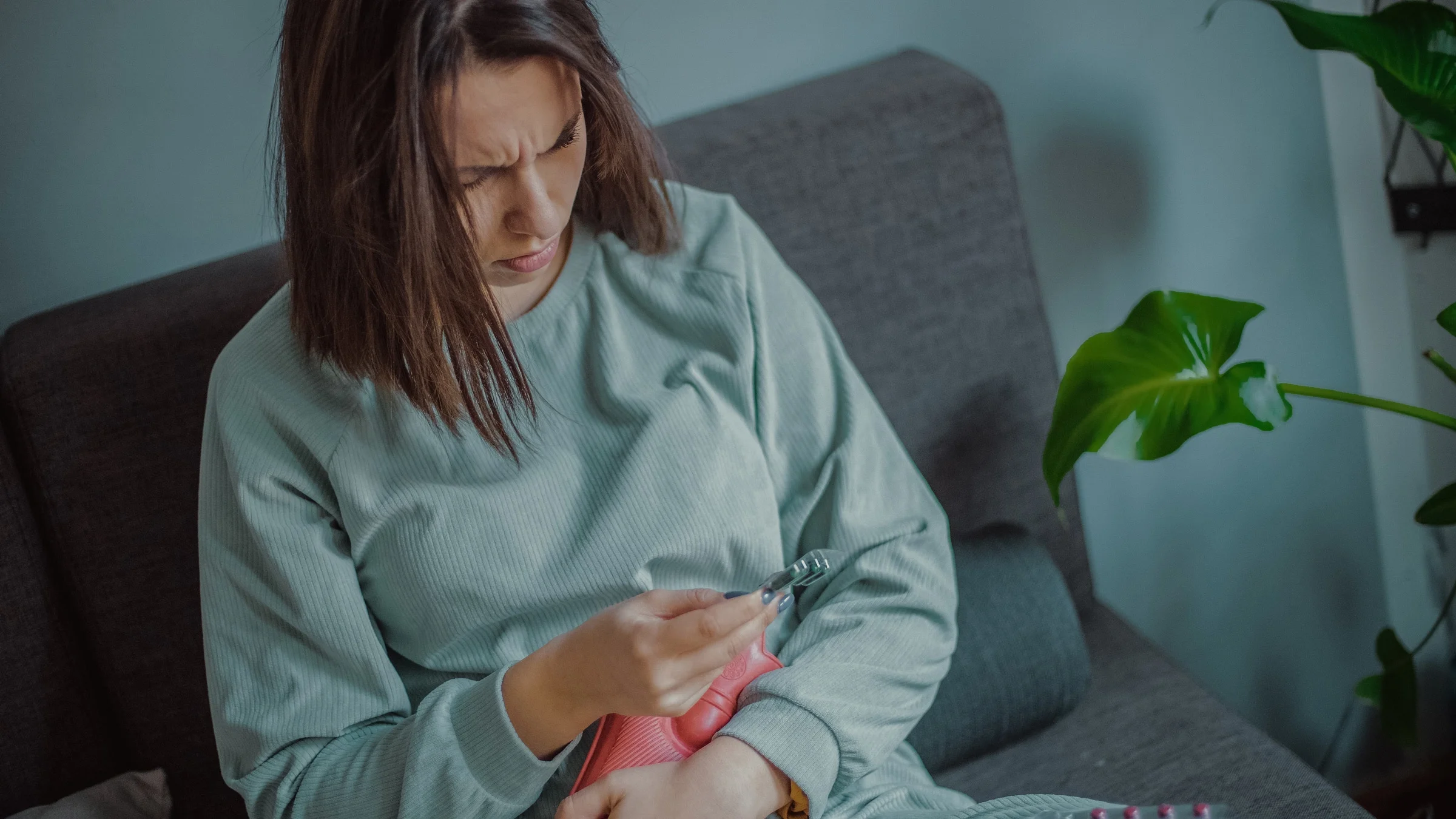 A person in pain on their couch, holding pills and a hot water bottle.