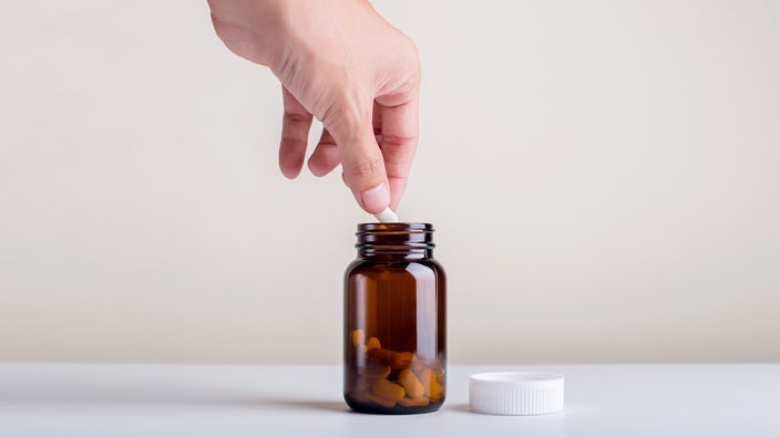 Glass pill bottle on a white background. There is a person's hand reaching in and grabbing a pill out of the bottle.