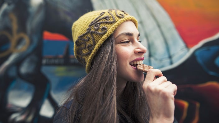 Woman in beanie eating a chocolate bar. She is standing on the street in front of a colorful mural.