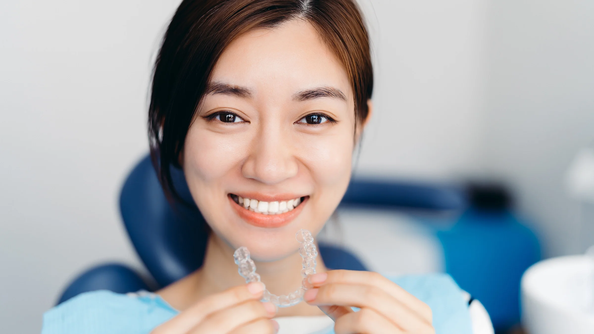 Close-up portrait of a young woman putting in a clear retainer. She is sitting in the dentist chair in the exam room.