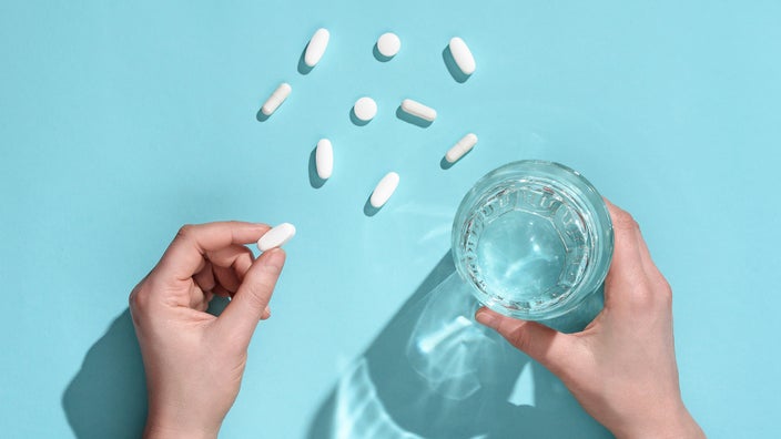 Top view of a hand holding pill and glass of water with extra pills on a blue surface.
