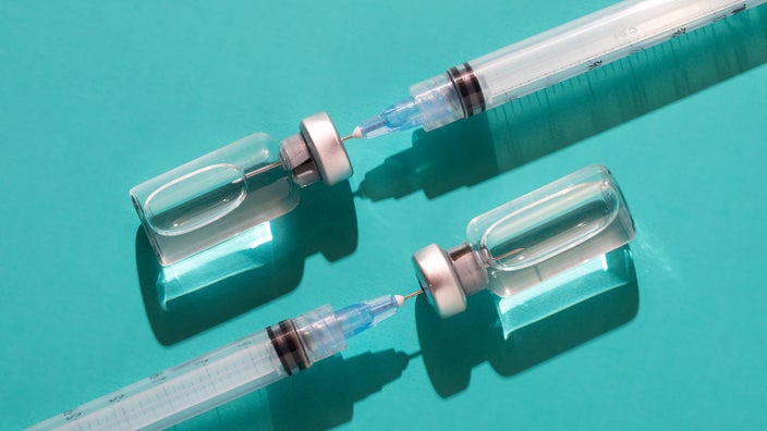 Two insulin vials and syringes on a teal colored background. The syringes are already stuck into the vials.