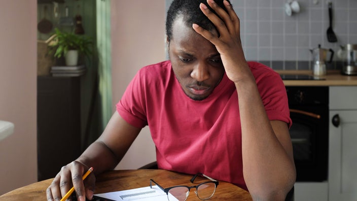 A man is looking anxious and holding his head in his hand while doing financial paperwork.