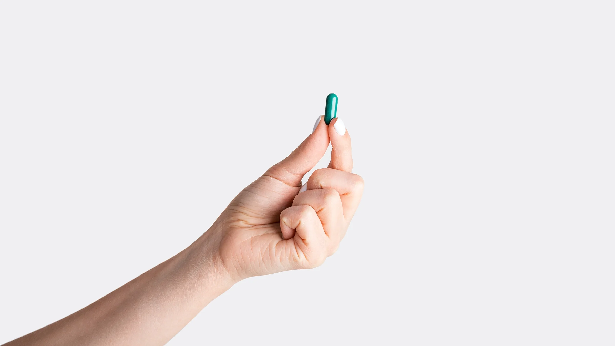 Person holding one green pill in the center of the frame. We can only see their arm reaching in holding the pill. All on a plain white studio background.