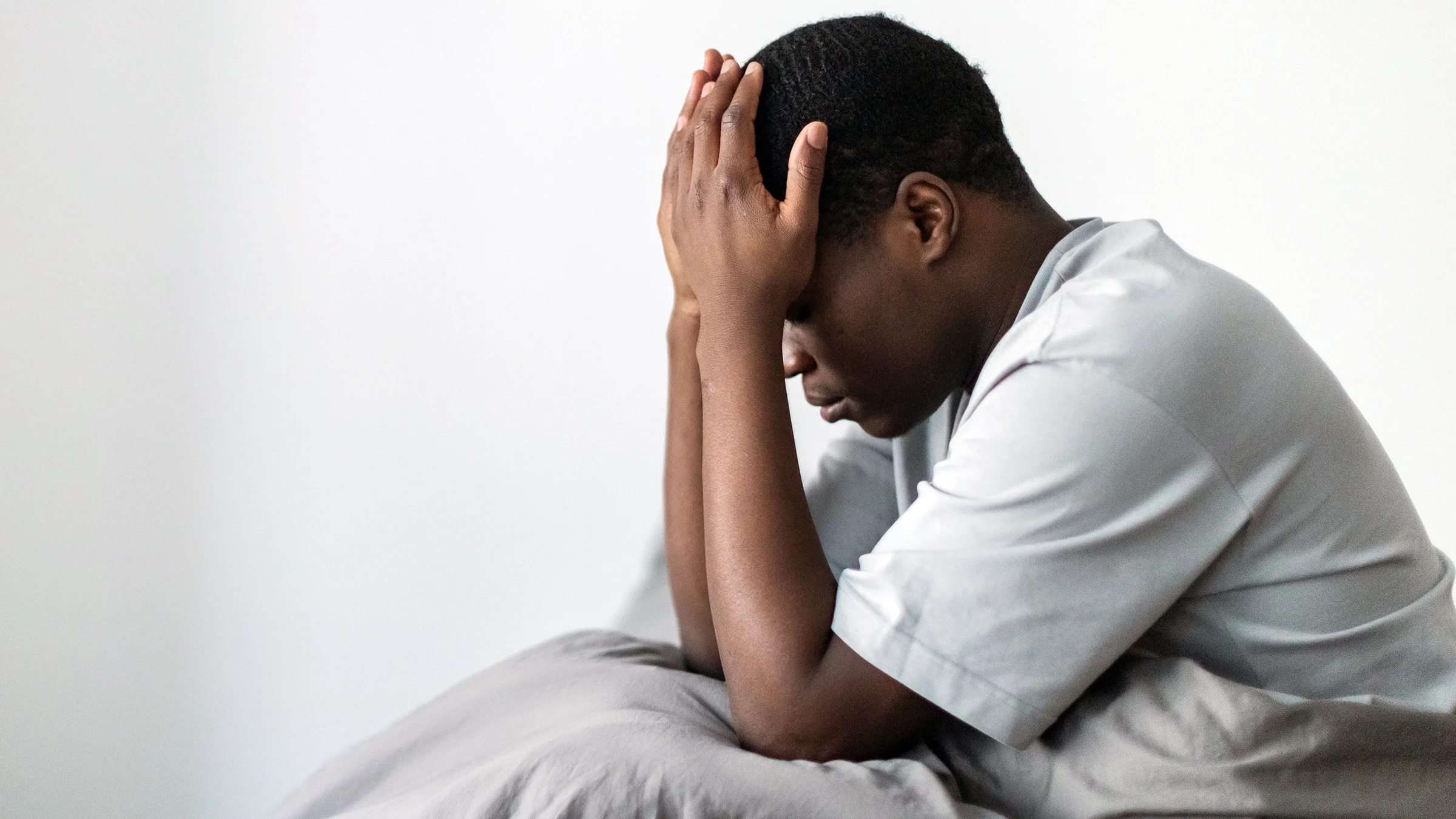 A man sits in bed while holding his head in his hands.