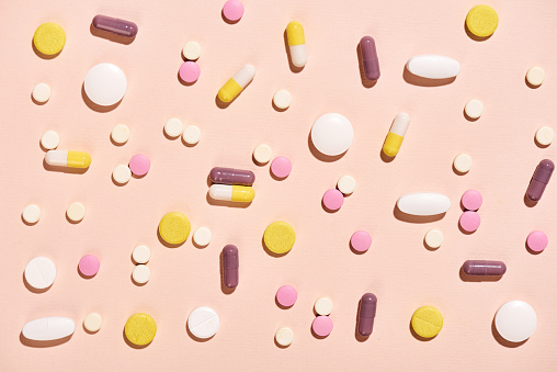 Various pills of different sizes and colors, some white, yellow, pink, and dark red, on a light pink background.