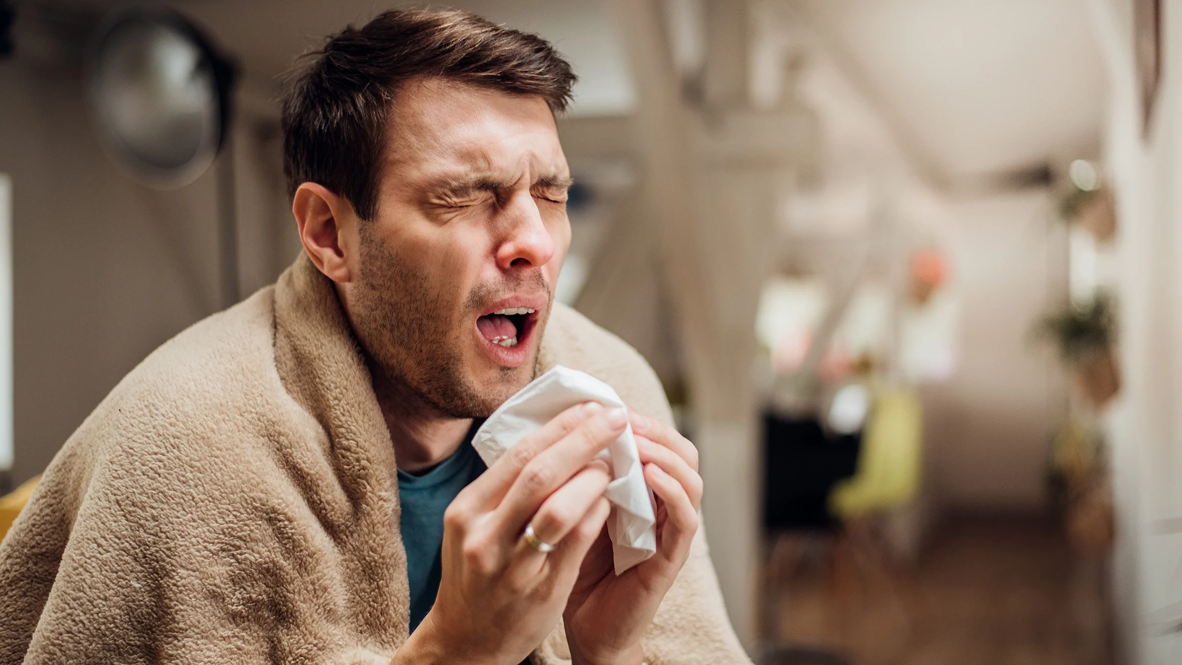 Man with blanket draped over his shoulders sneezing while he is sick at home.