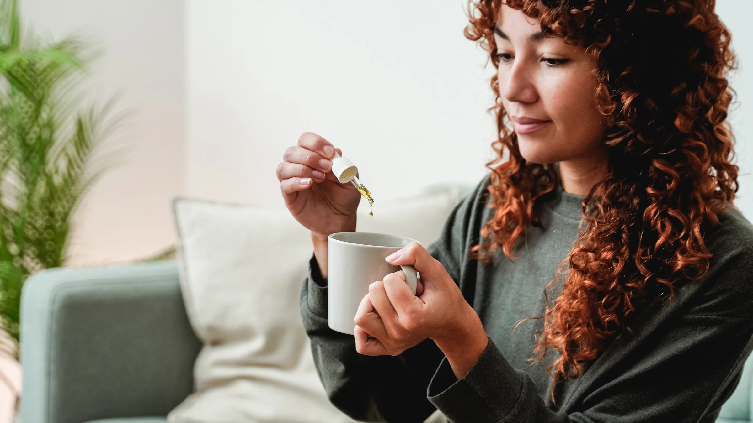 Woman sitting at home on her couch putting CBD oil drops into her cup of tea.