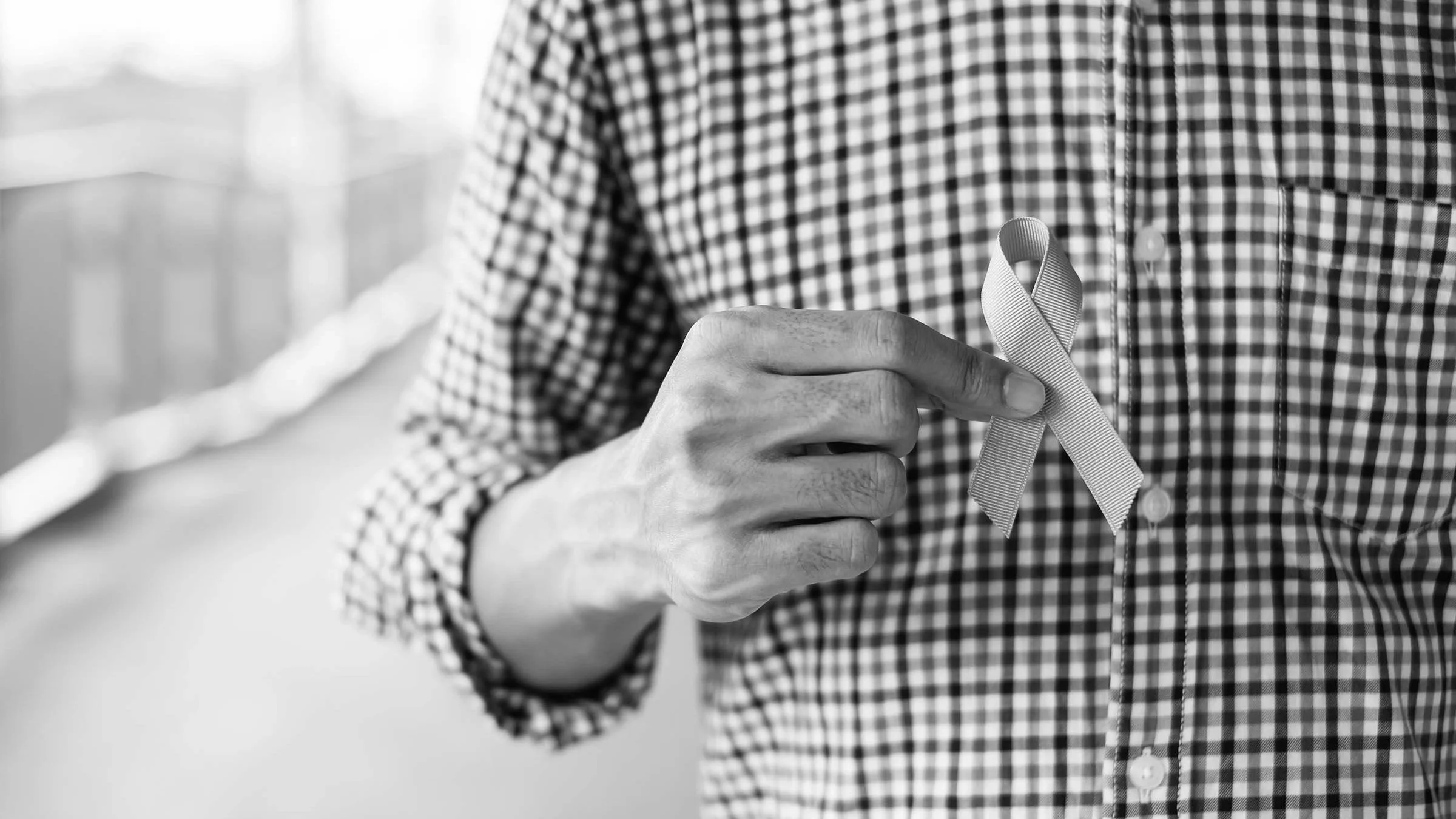 Black and white cropped image of a man holding cause ribbon in front of his chest. He is wearing a gingham button up shirt.