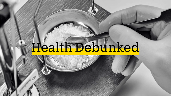 Black and white image of a gloved hand weighing out white powder that is artificial sweetener in a precision scale. There is added text on top reading “Health Debunked” with a yellow highlight bar behind it.