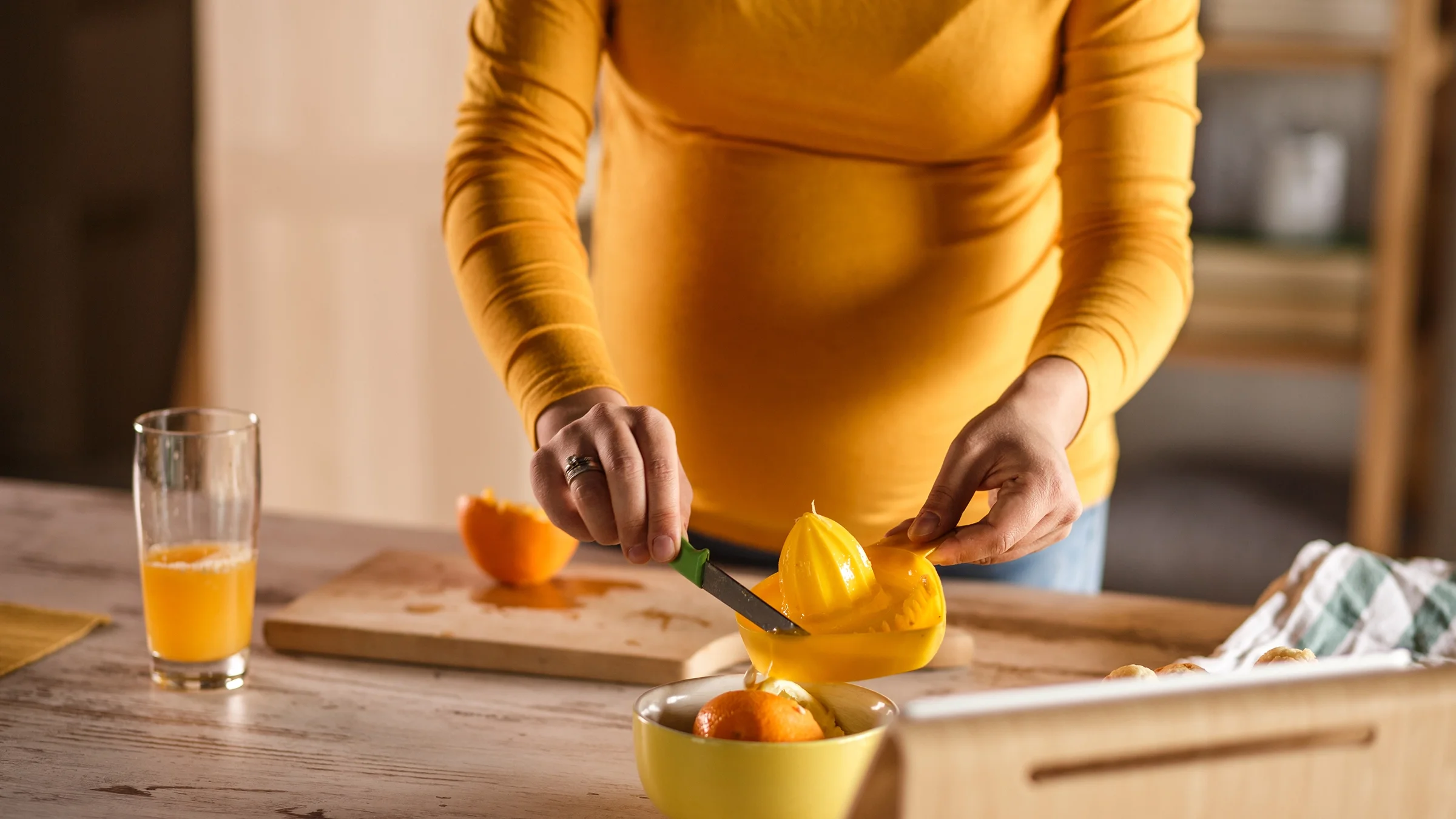 Cropped shot of a pregnant person making orange juice. They’re wearing a yellow-orange long-sleeve shirt.