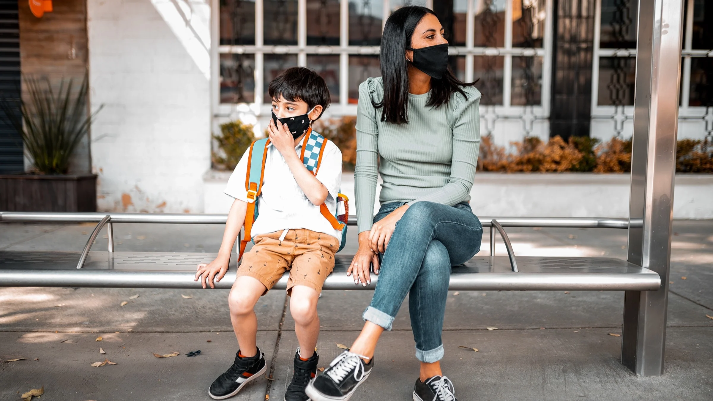 A young mom and son sitting on a bus bench wearing face masks looking either direction for the coming bus.