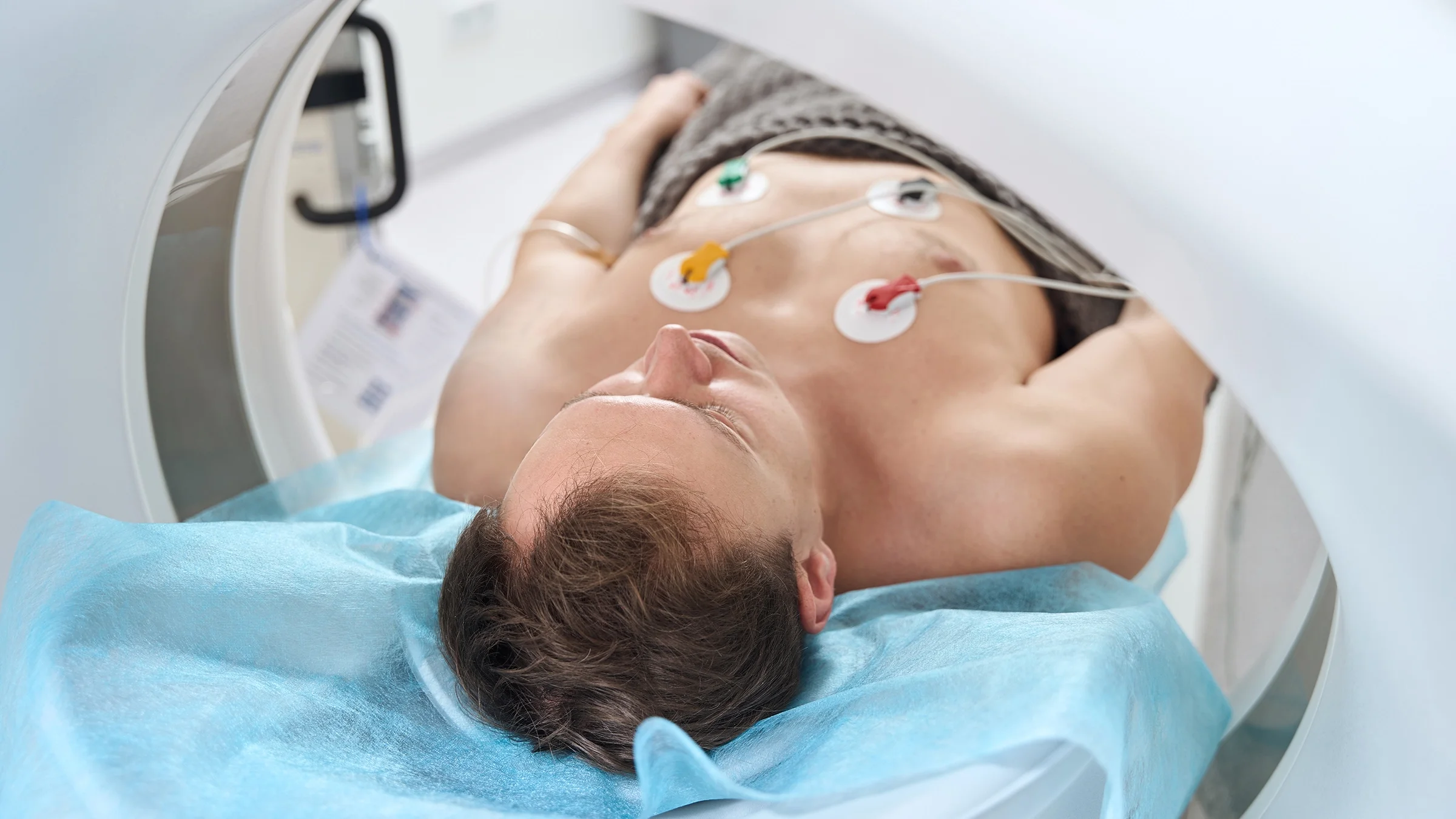 A man with ECG electrodes attached to his chest is lying inside a CT scan machine.