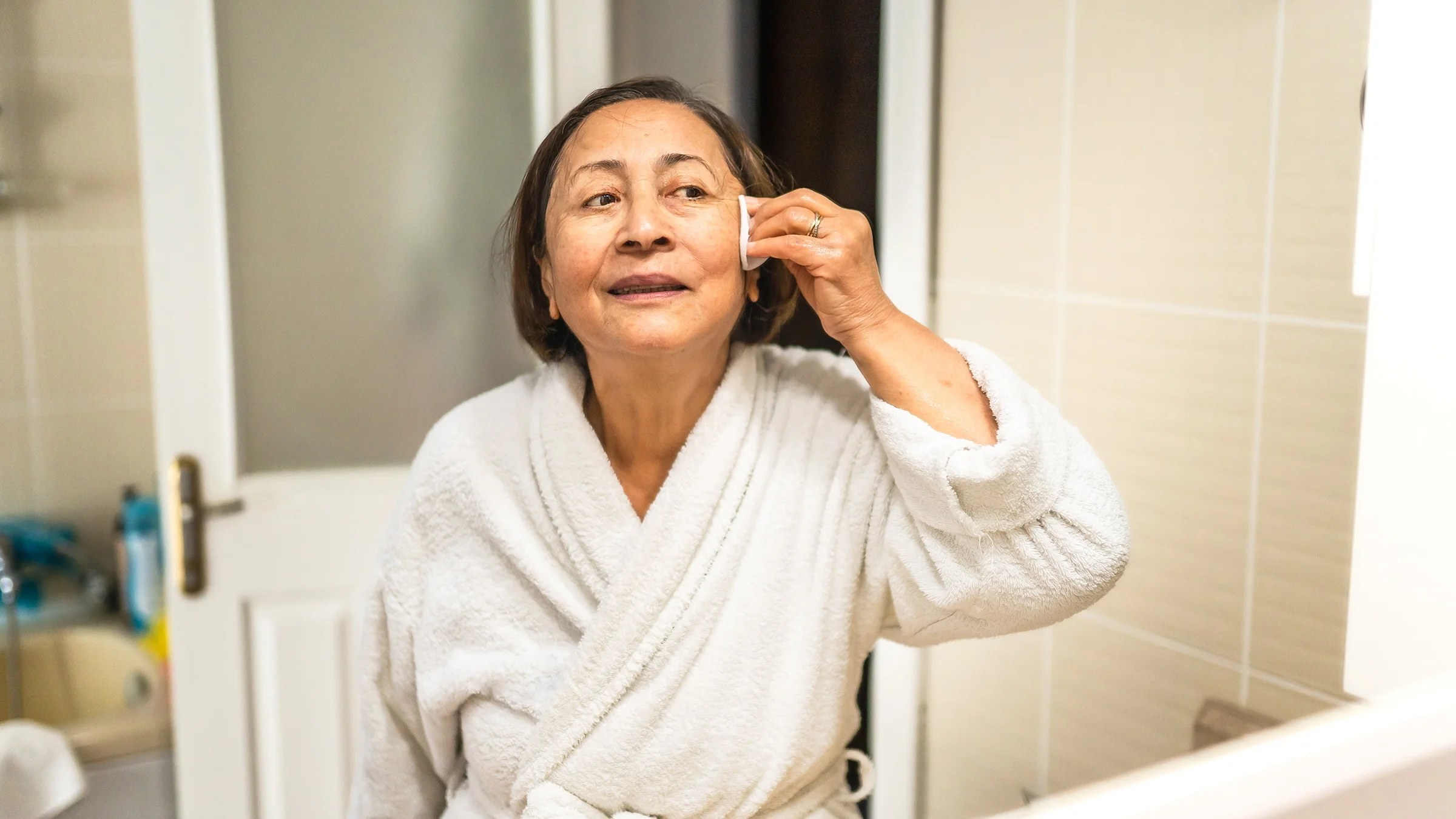 Older woman doing her skin care routine in the bathroom mirror.