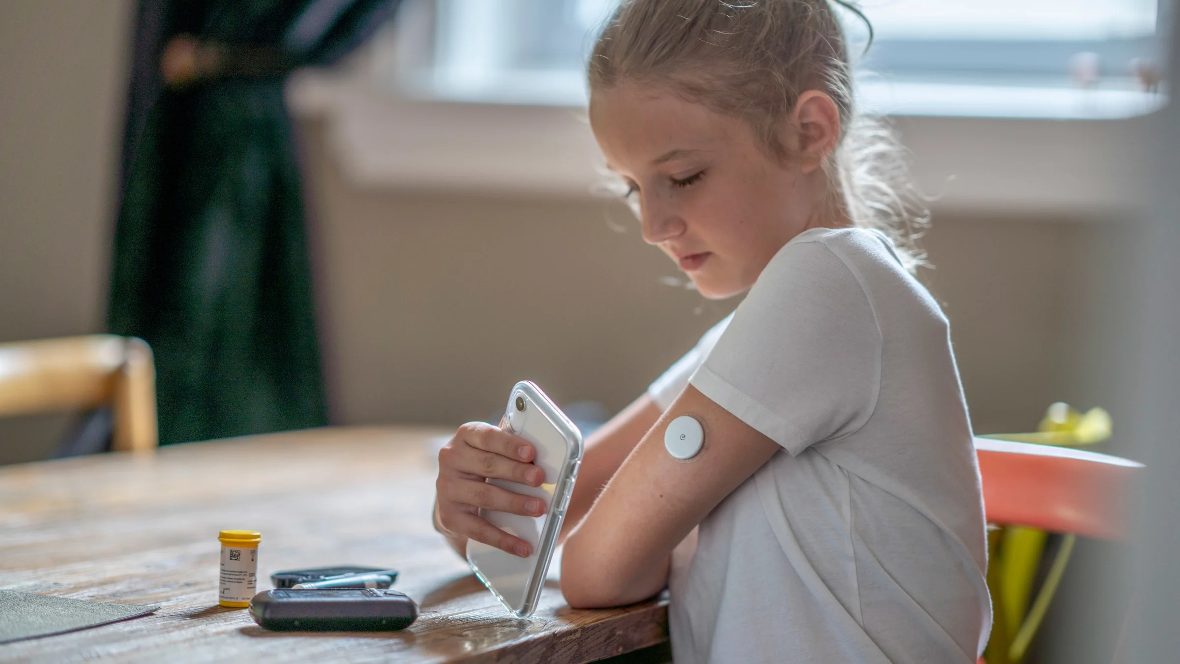A teen using a glucometer. There is a pill bottle on the dining table.