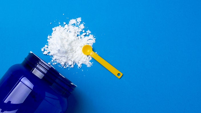 White creatine powder is spilling out of a blue supplement jar on a blue background. There is a yellow scoop on top of the powder.