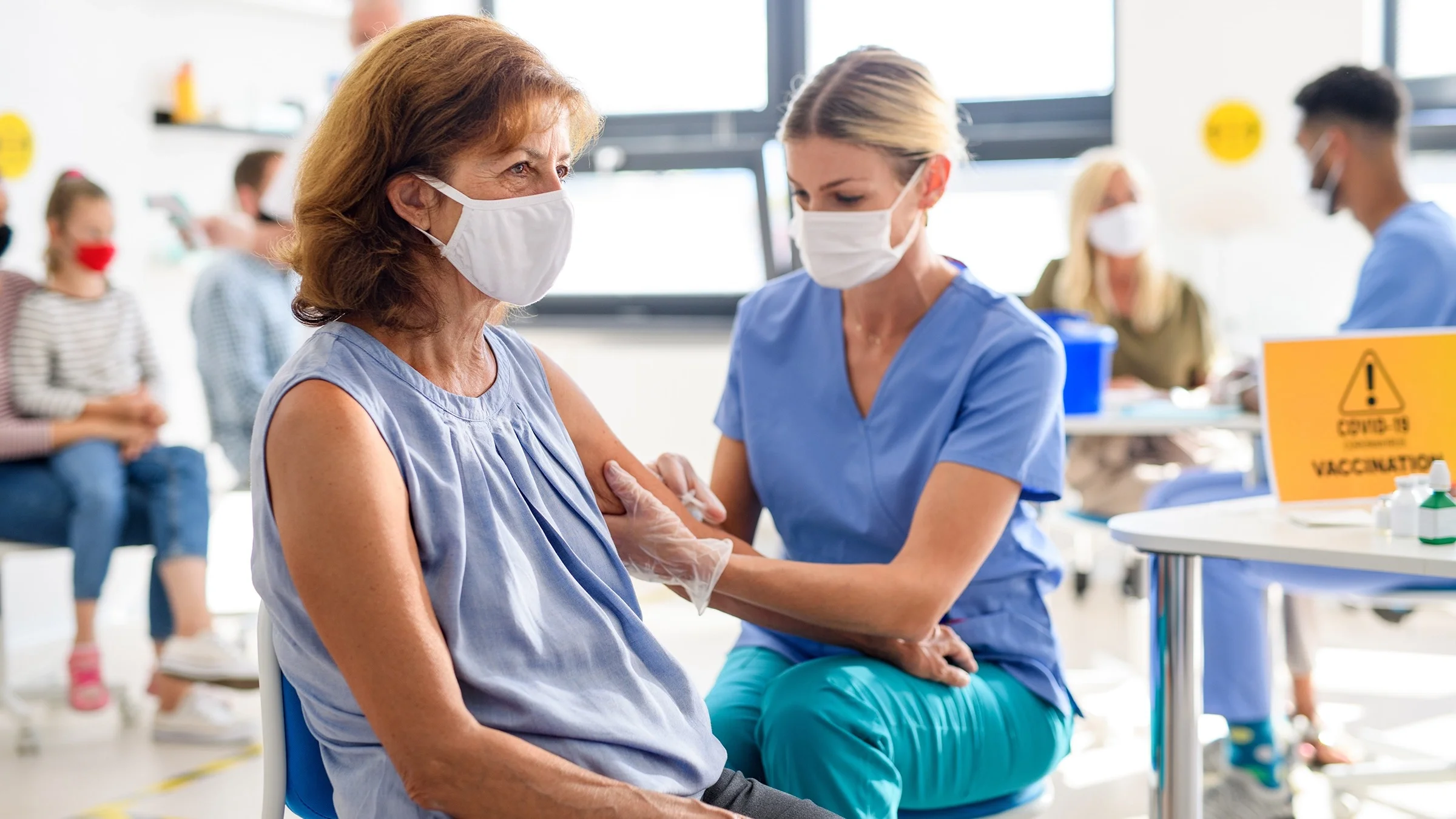 Older woman with face mask getting her COVID-19 vaccine at a community vaccination site. The nurse is administering the shot while the woman looks off into the distance.