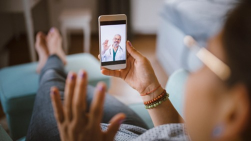 Close-up of a woman on a telehealth visit on her phone waving hit–the perspective is over her head and should looking at the phone screen.