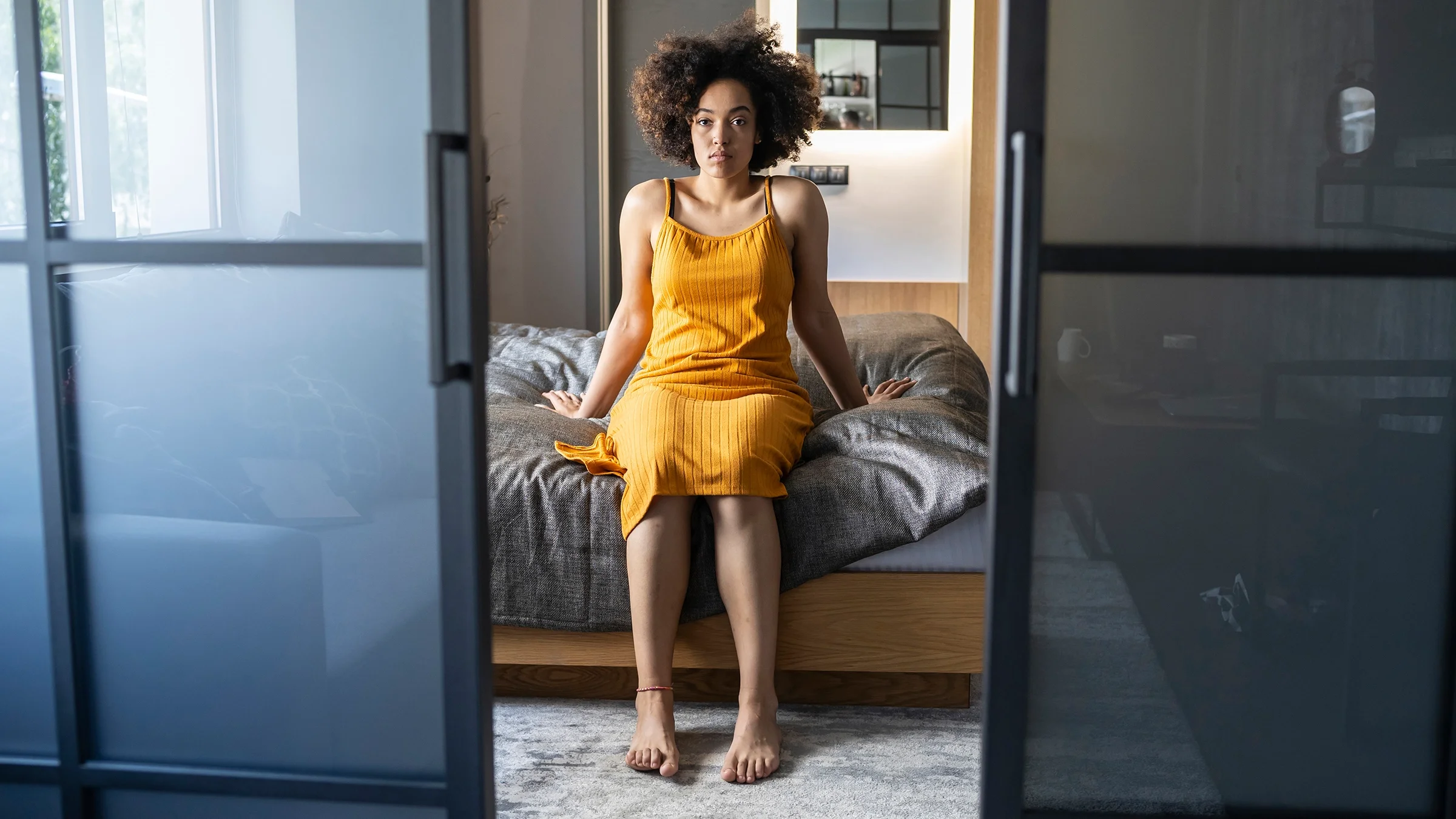 Woman sitting on the edge of the bed looking at the camera. The camera is positioned from the outside looking in. You can see the doorway and doors open in front of the woman.