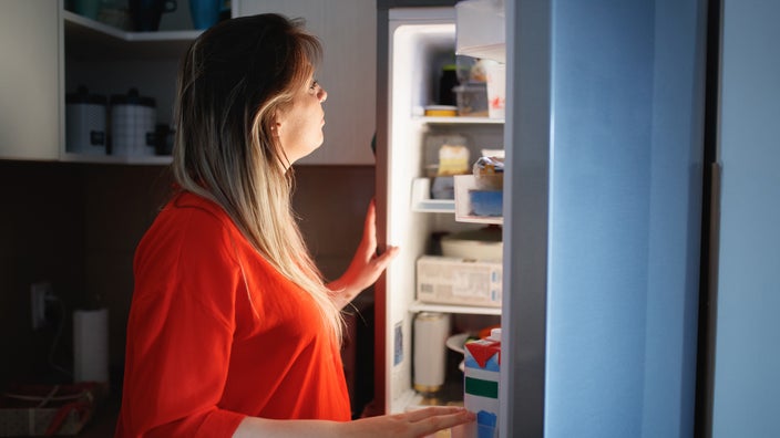 Blonde woman looking in her fridge for something to eat. Most of the light in the frame is coming from inside the fridge.