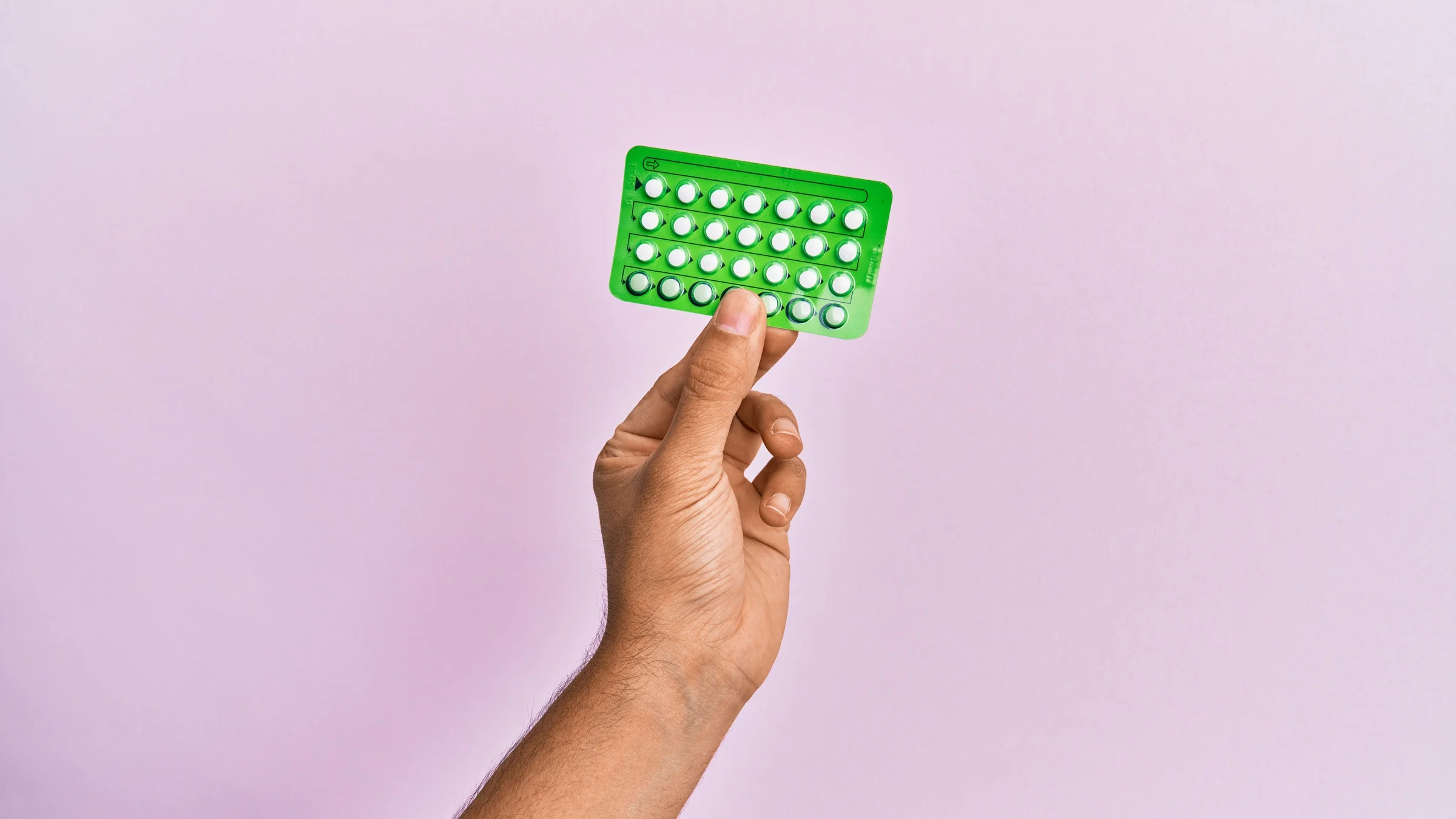 Tan hand holding a green pack of birth control pills against a lilac background.