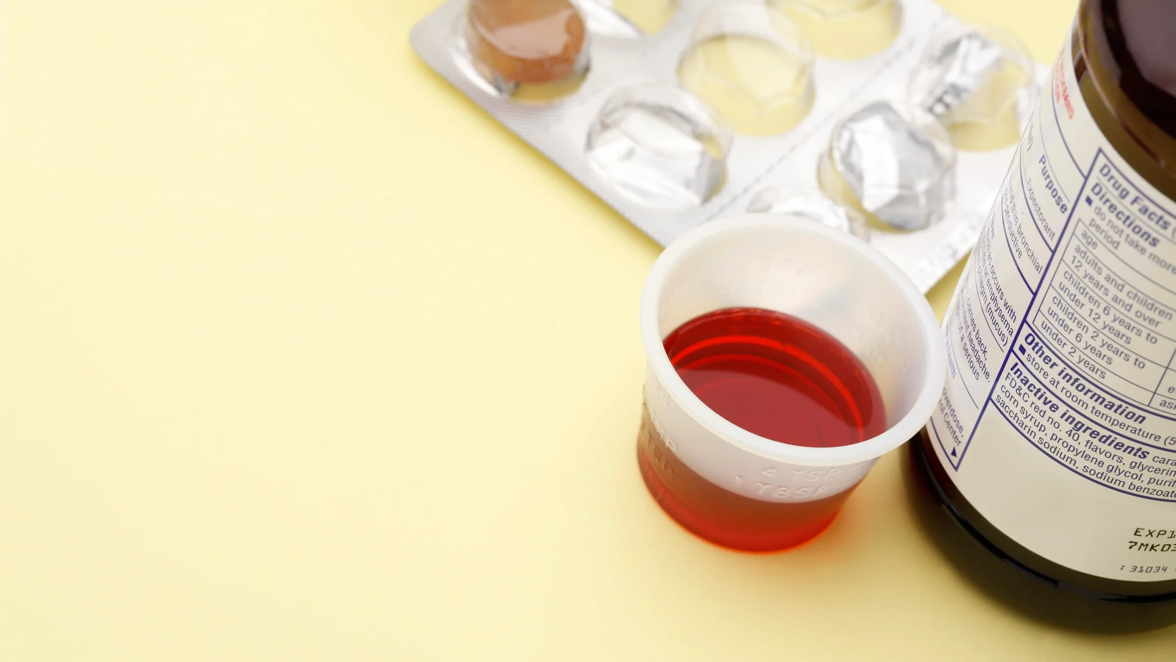 A liquid medicine cup and cough drop package sit before a yellow background.