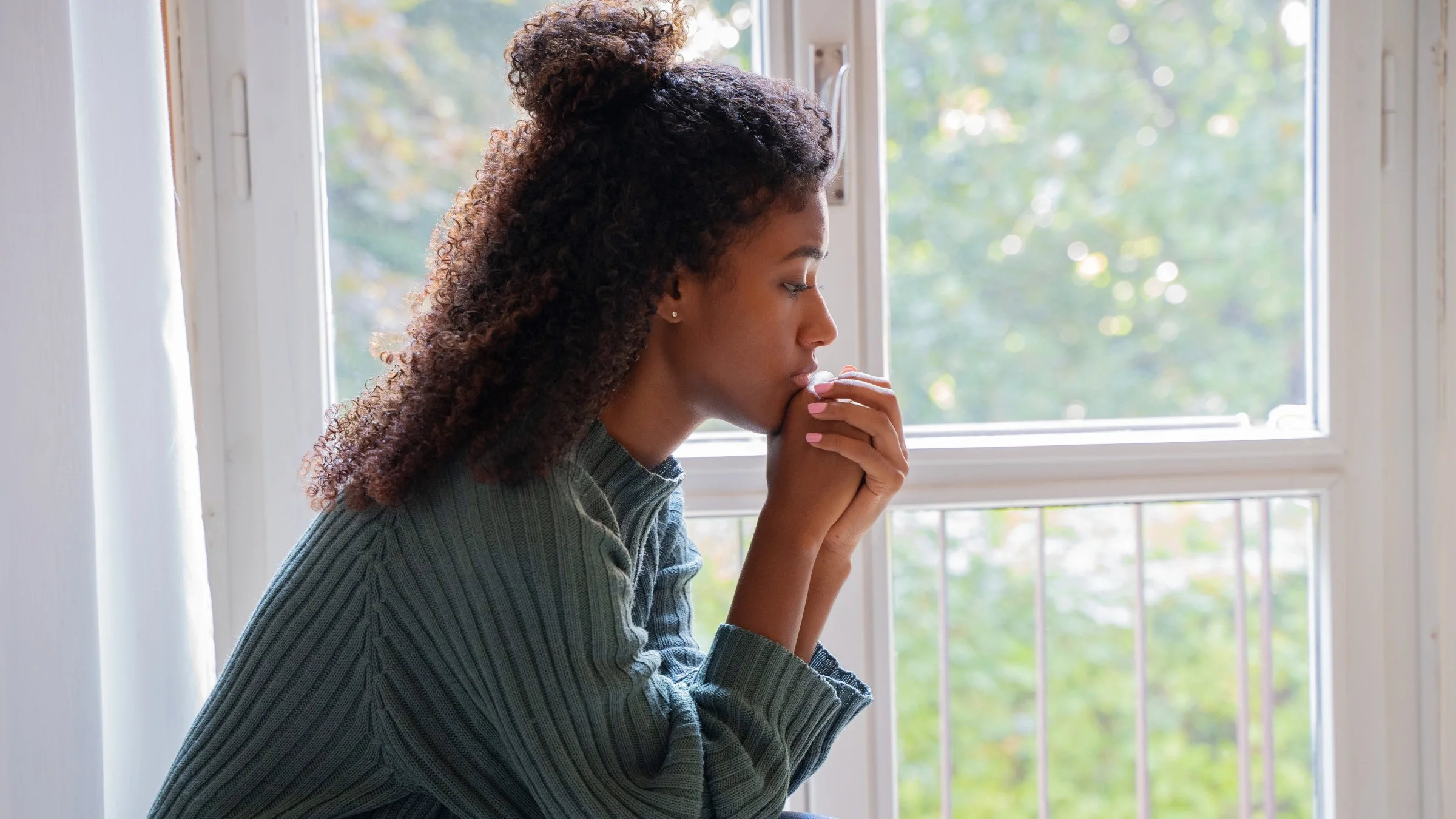Young woman in a green sweater with curly hair sitting at a window looking depressed.