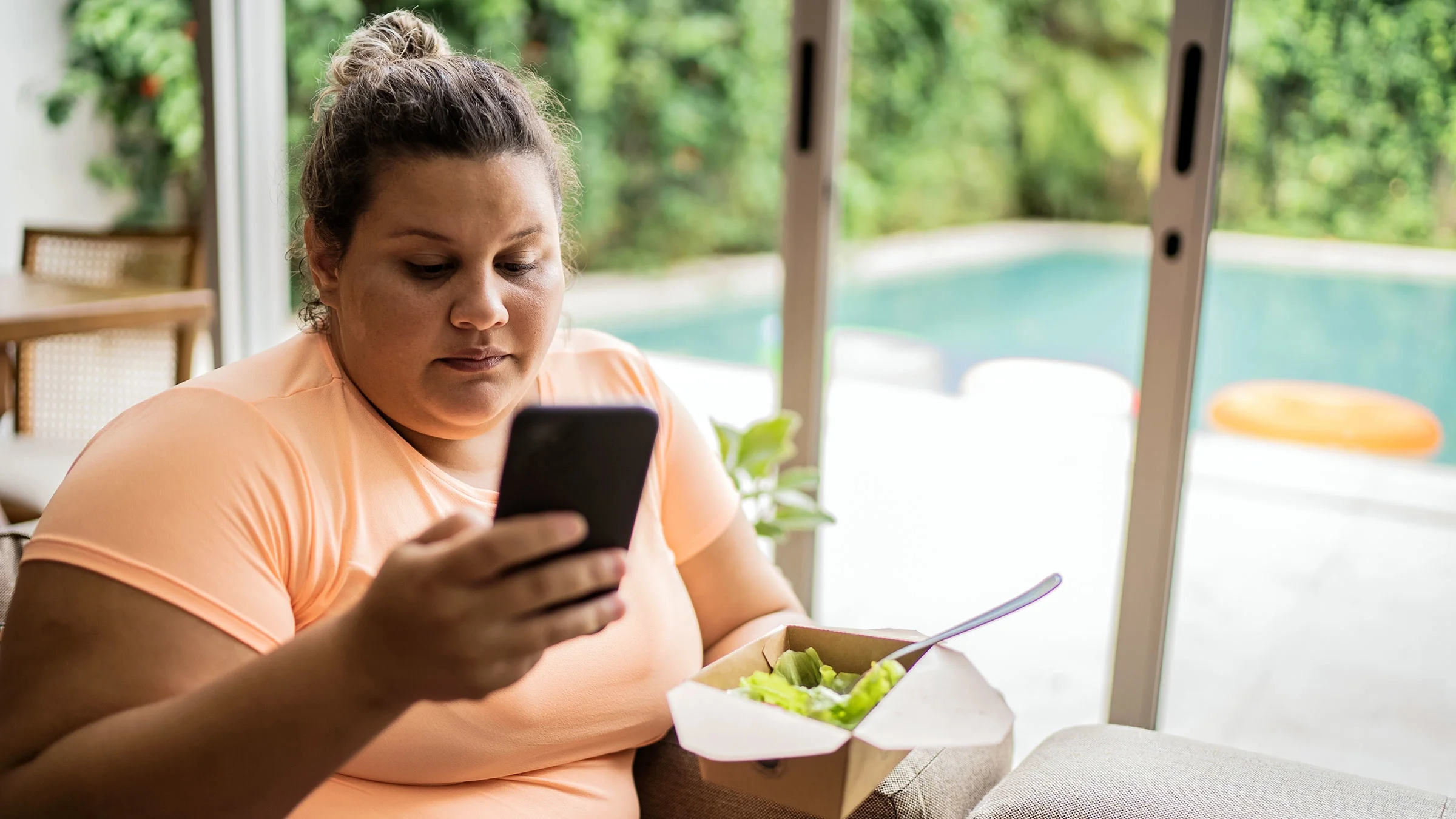 A woman looks at her phone while eating a salad.