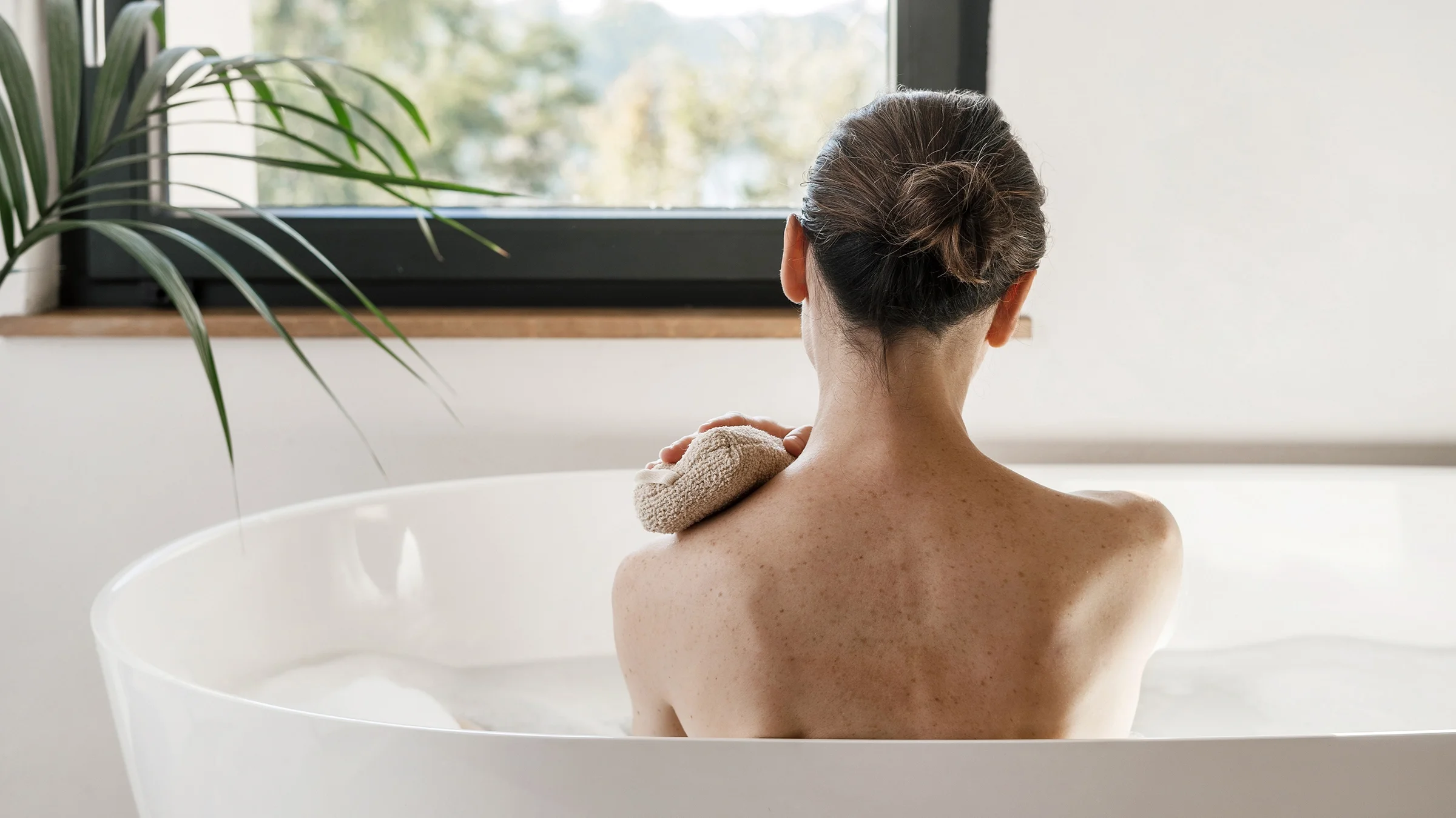 Rearview shot of a woman washing her body with a sponge during a bath.