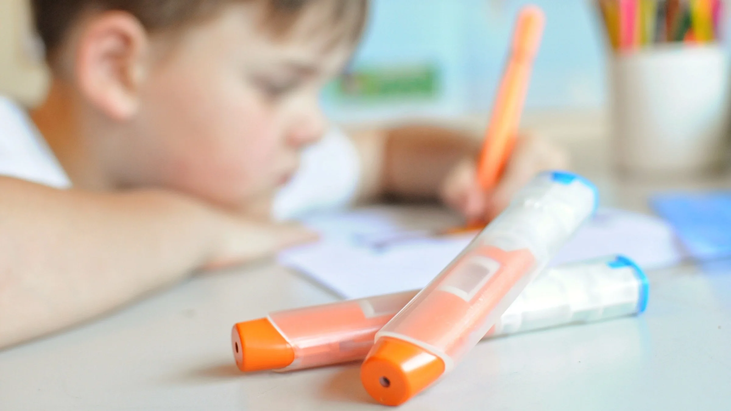 Young child out of focus drawing at his school desk with anaphylaxis injector pens next to him.