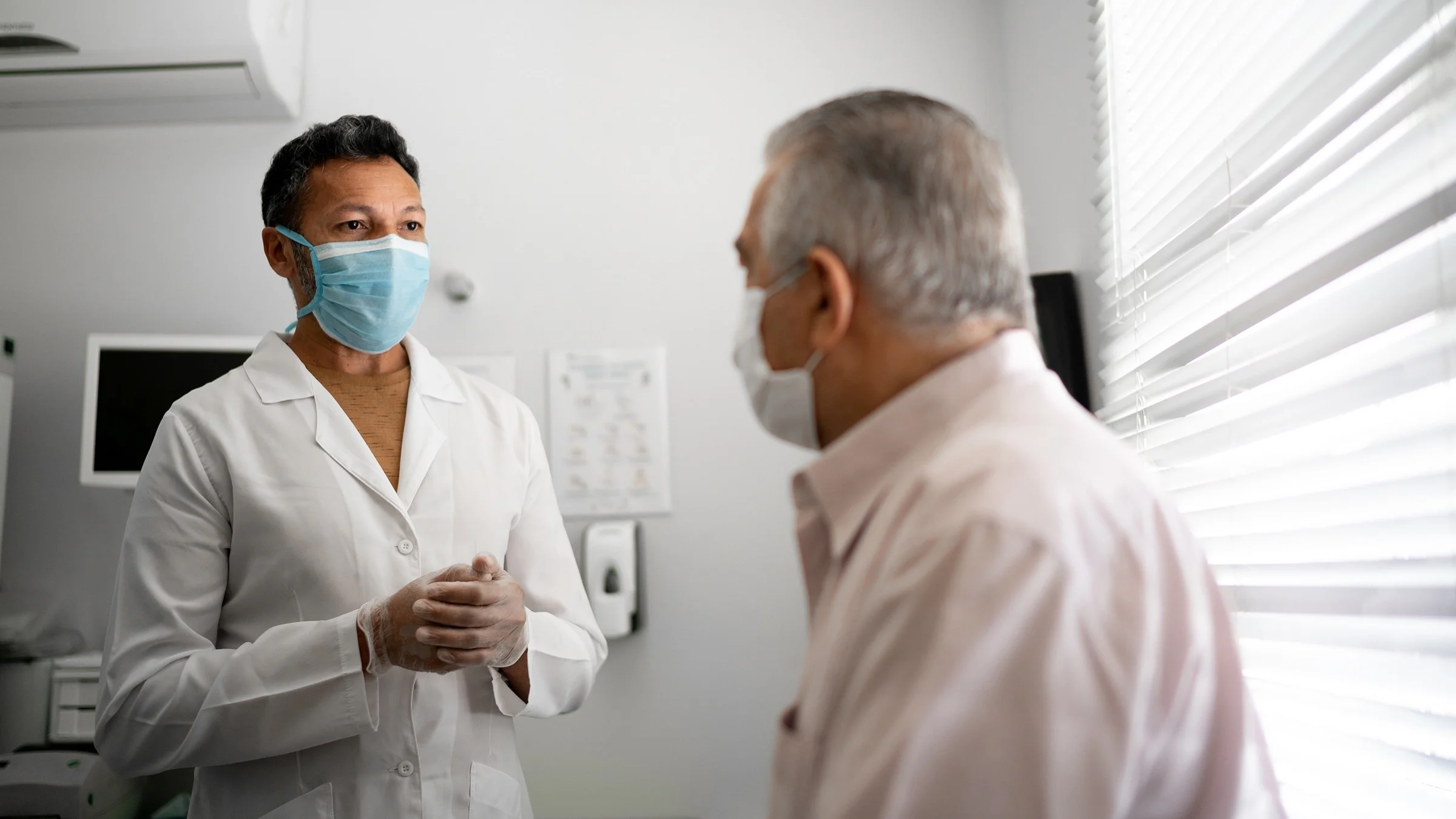 Older male patient with his doctor in the exam room. The doctor has a blue medical face mask on and clear medical gloves. Both have neutral looks on their faces.