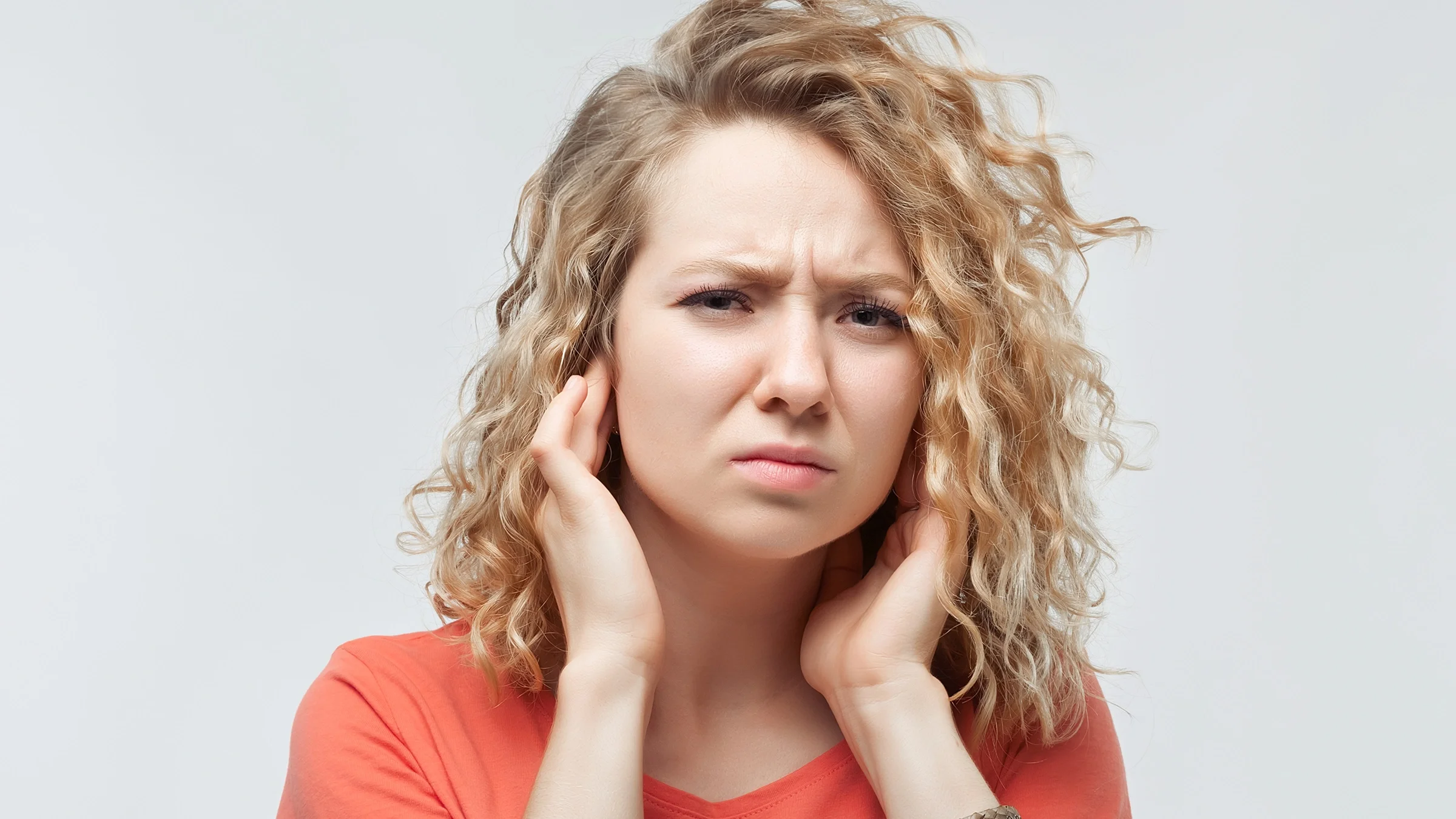 Portrait of a woman with blond, curly hair on a plain white background. She is holding her ears and furrowing her brow.
