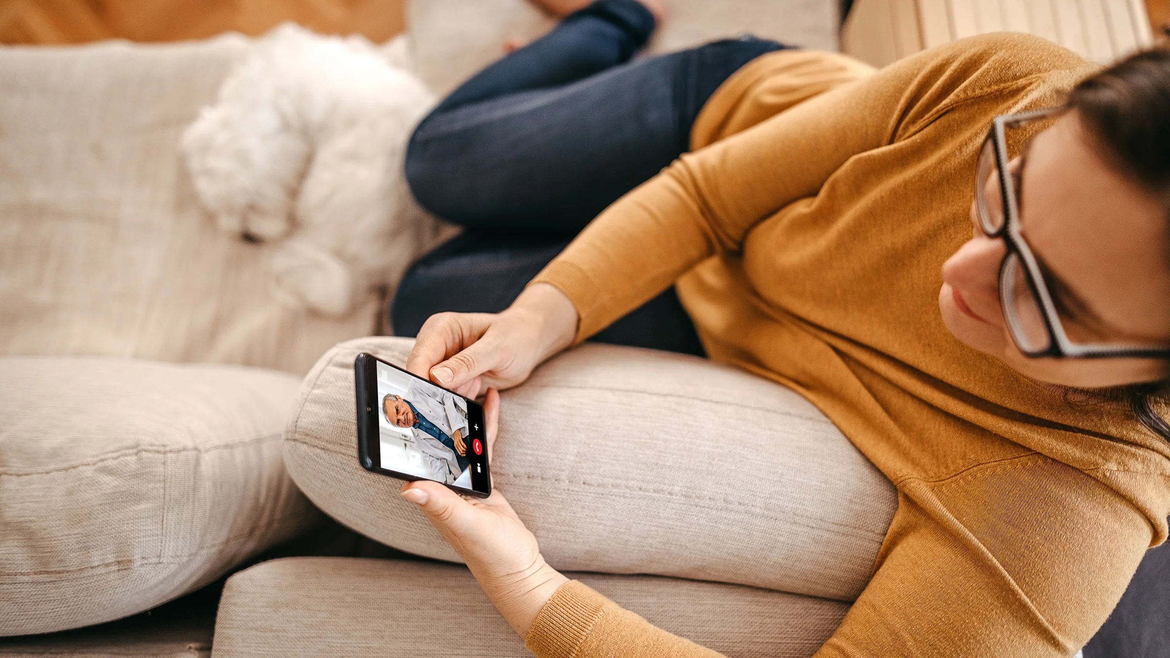 Overhead shot of a woman on her mobile phone for a telehealth visit with her doctor. She is sitting on her couch with her white fluffy dog curled up next to her.