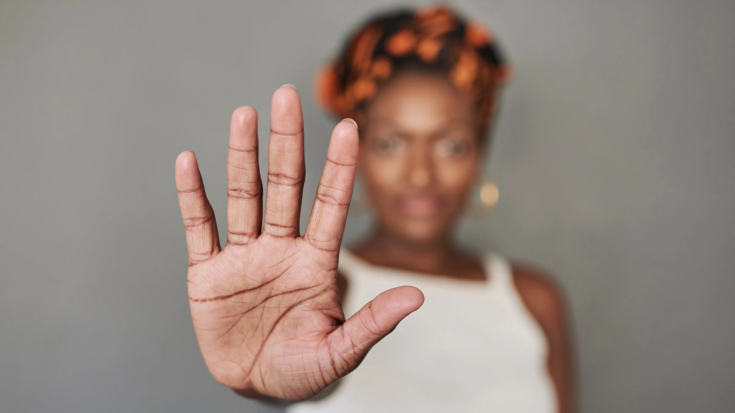 Portrait of a young Black woman against a gray background. Her hand is outstretched in a ‘stop’ motion while she is in the background, out of focus.