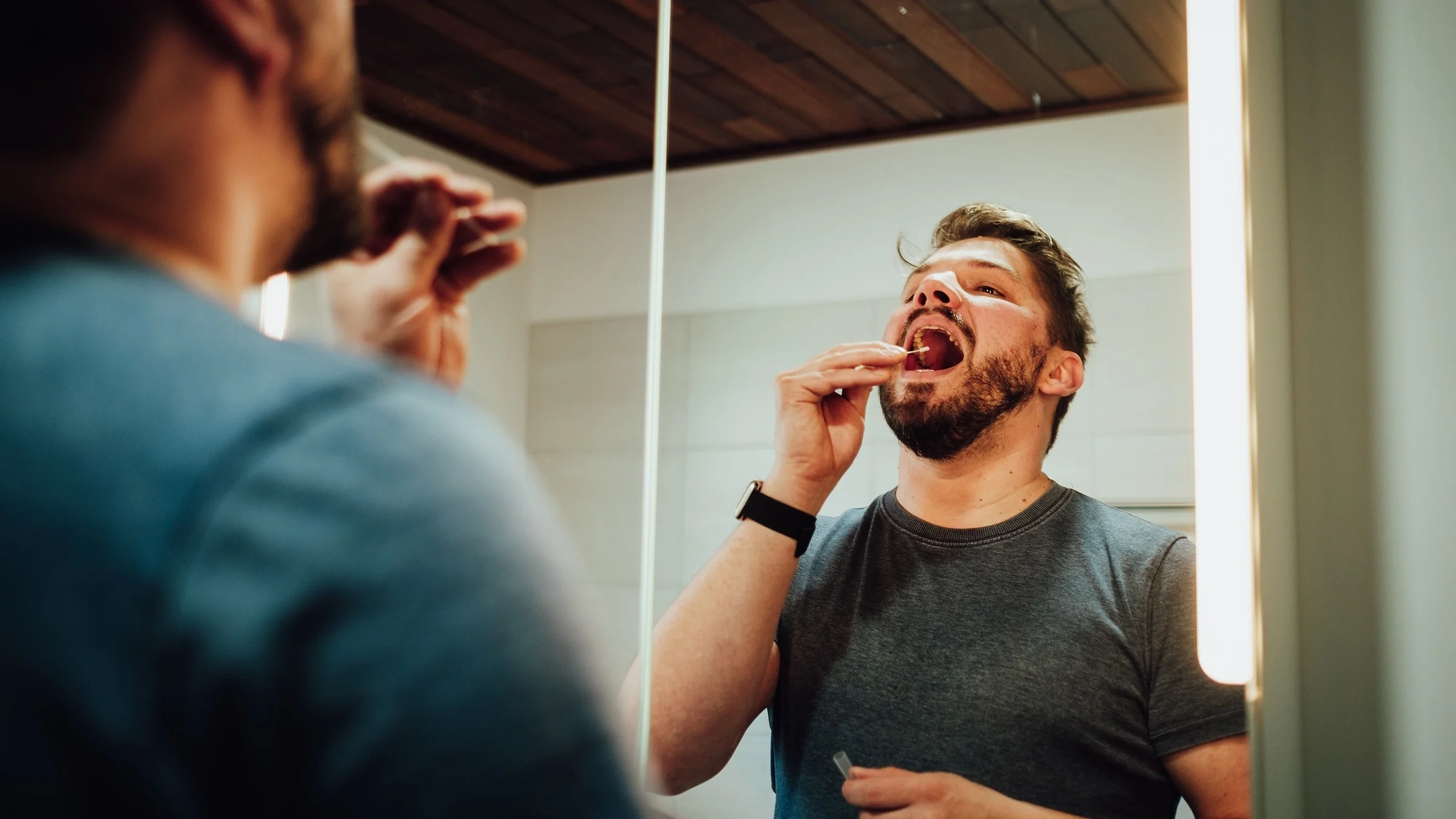 Man using an at home COVID test in his bathroom. He is using the swab on his throat and mouth. The photo is shot over his should and looking at him in the mirror.