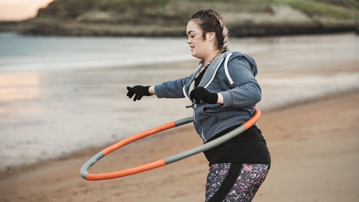 A woman is training on the beach with a weighted hula hoop.