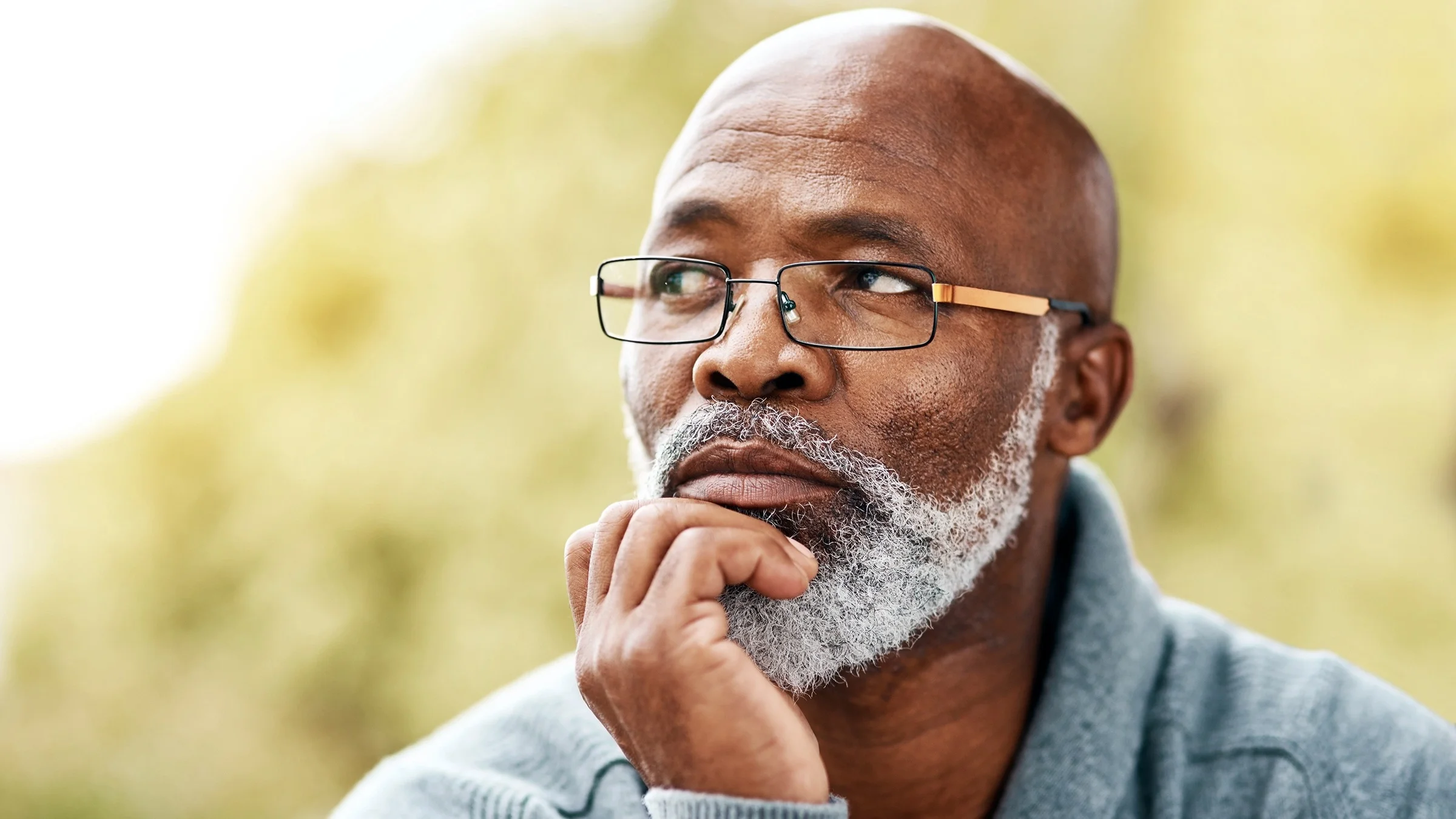 Portrait of a senior man with glasses looking off to the side. There is a bright yellow green garden background behind him.