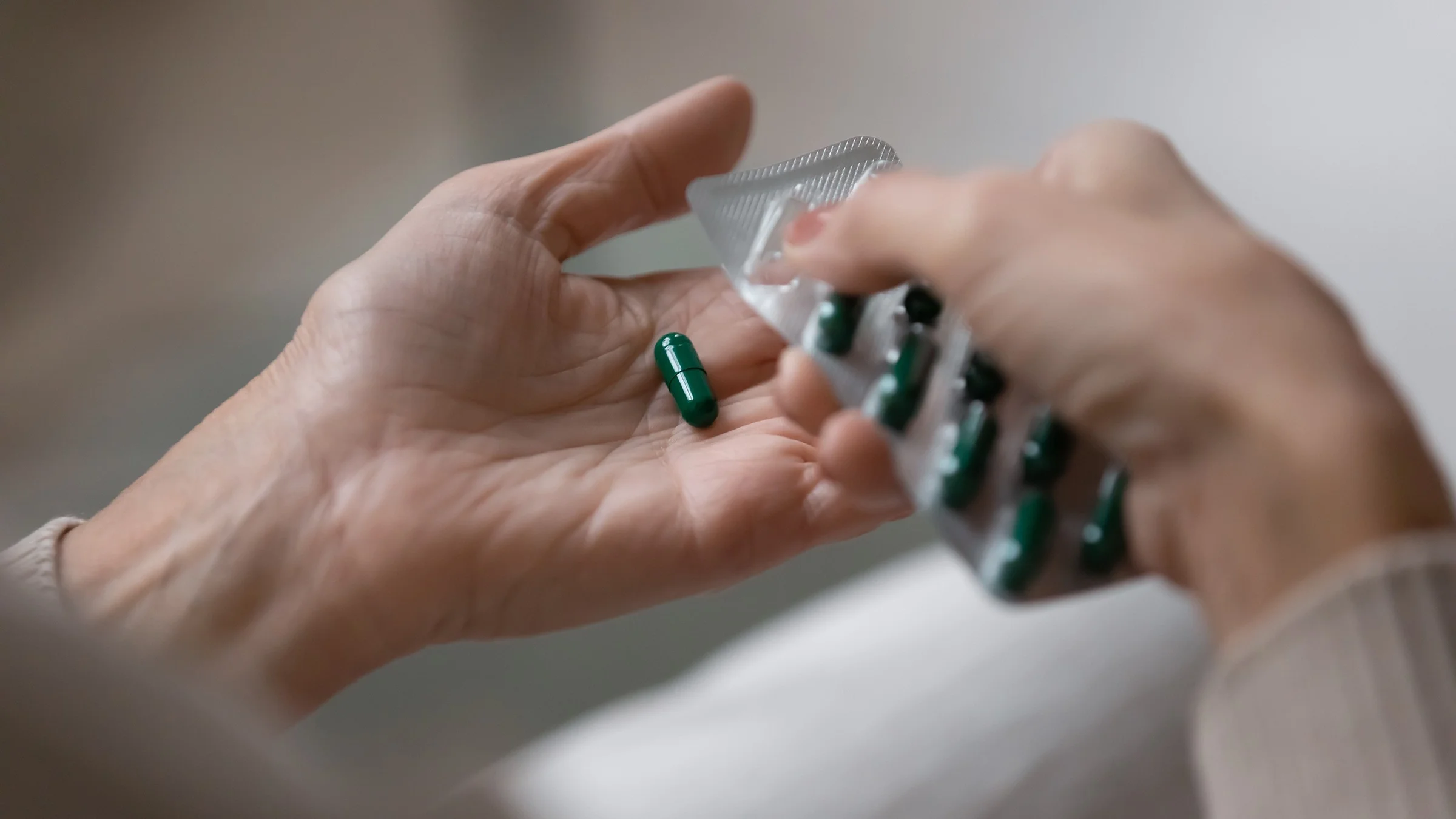 Close-up of a person taking a capsule pill out of a blister pack. The pills are dark green.