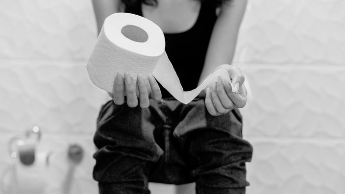 Black and white cropped shot of a woman sitting on a toilet holding a roll of toilet paper in her hands. She is wearing jeans and a tank top.