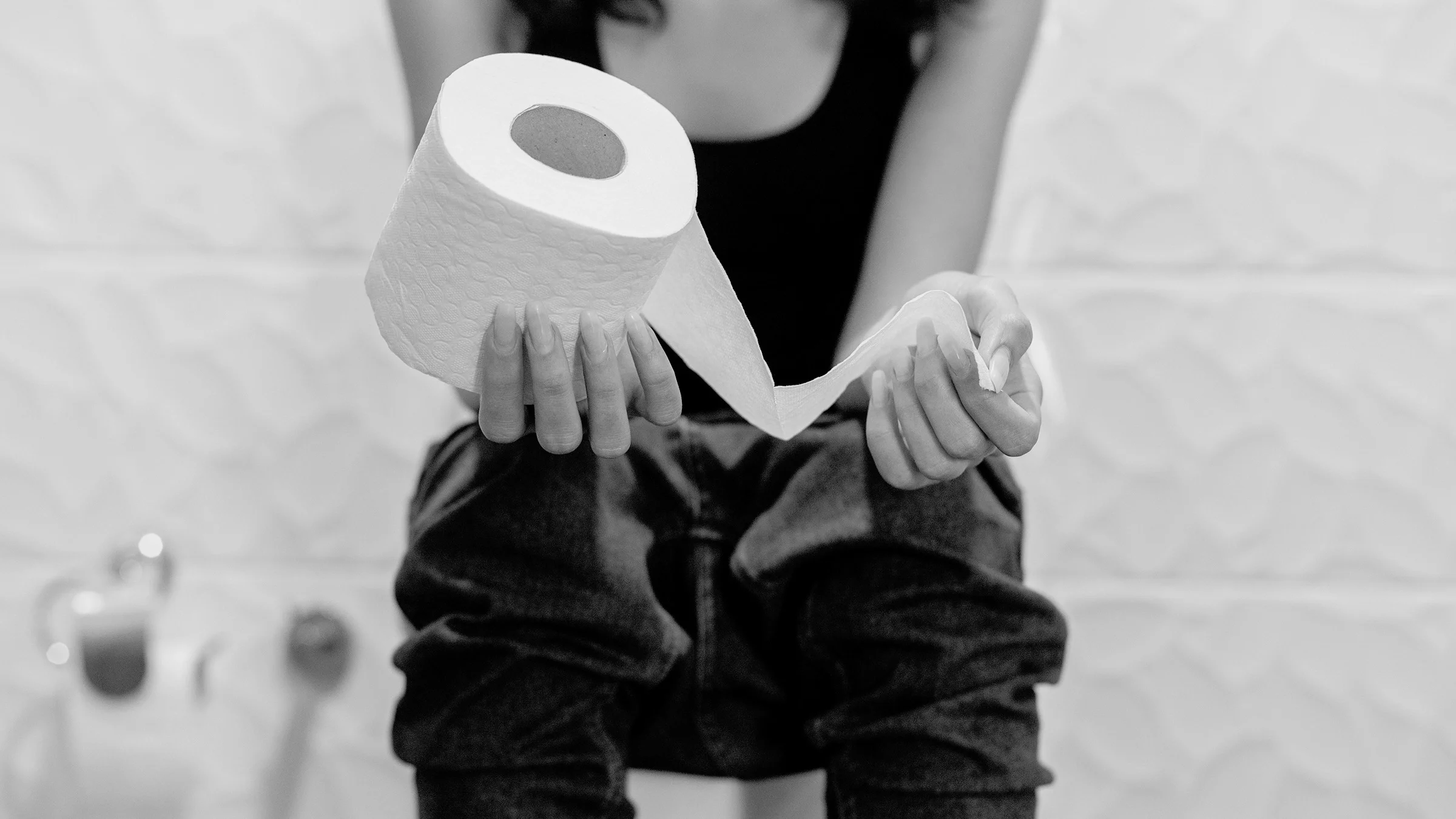 Black and white cropped shot of a woman sitting on a toilet holding a roll of toilet paper in her hands. She is wearing jeans and a tank top.