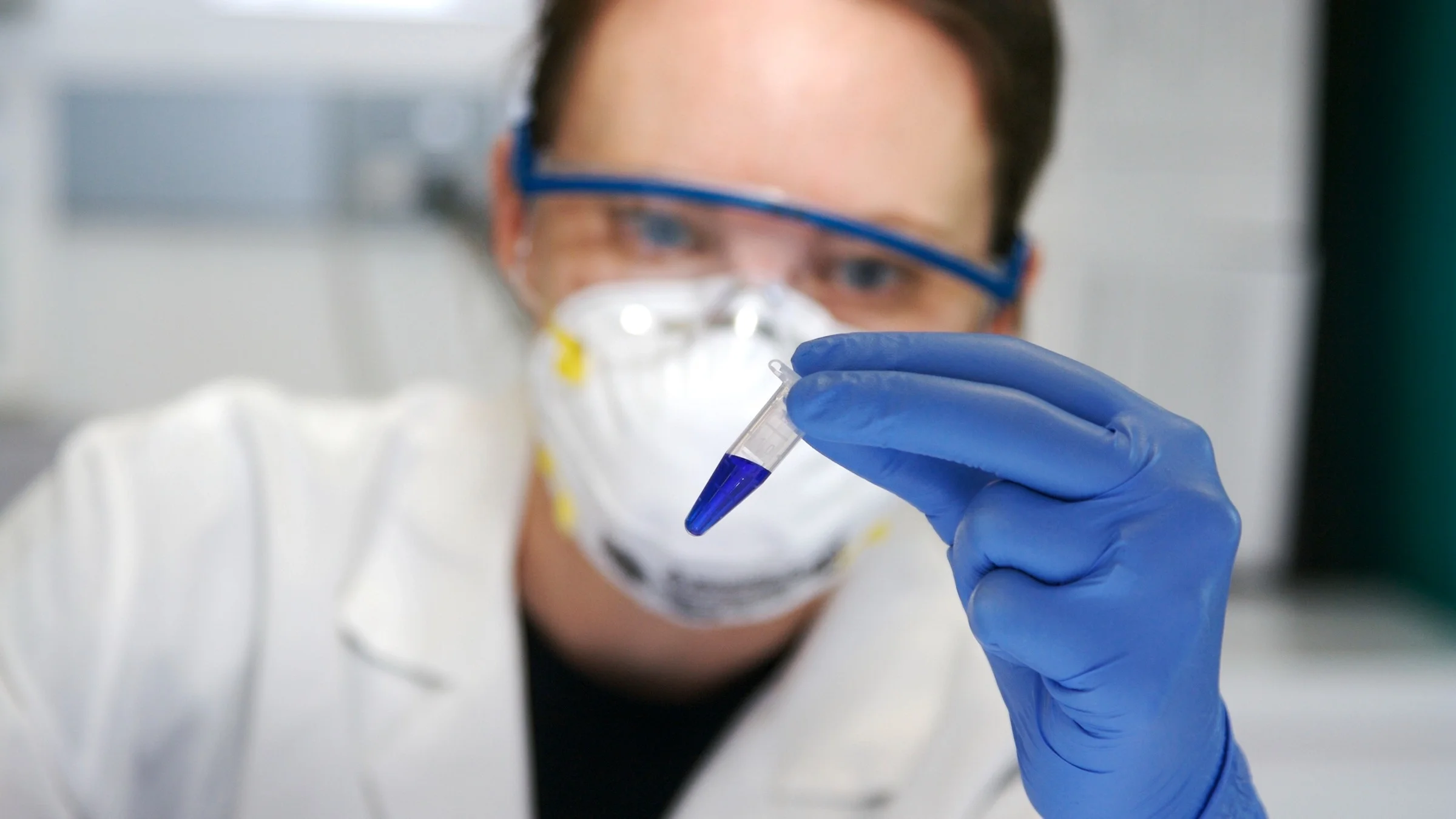 Close-up on a woman in the lab looking at blue liquid in a small test tube. She has on protective goggles and a N95 mask. She is blurry in the background while the test tube and her hand are in focus.