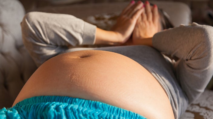 Cropped shot of a pregnant woman laying down on her back. The focus is on her exposed belly but you can see her in the background blurry. She has her hands over her face in exasperation and stress.