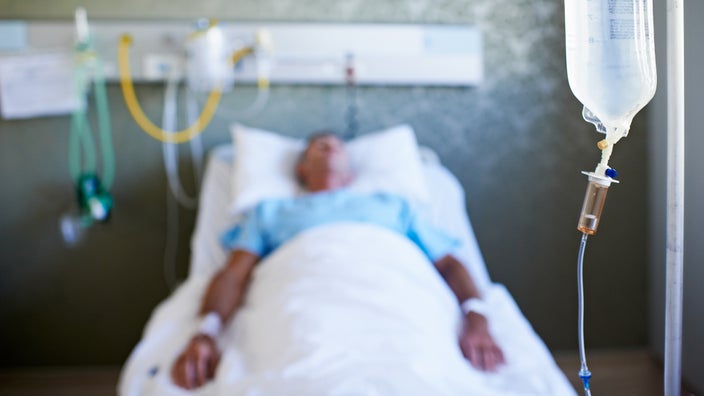An unconscious man lying in a hospital bed. He is in the background, which is blurry. The focus is on the IV bag to the right of the frame.