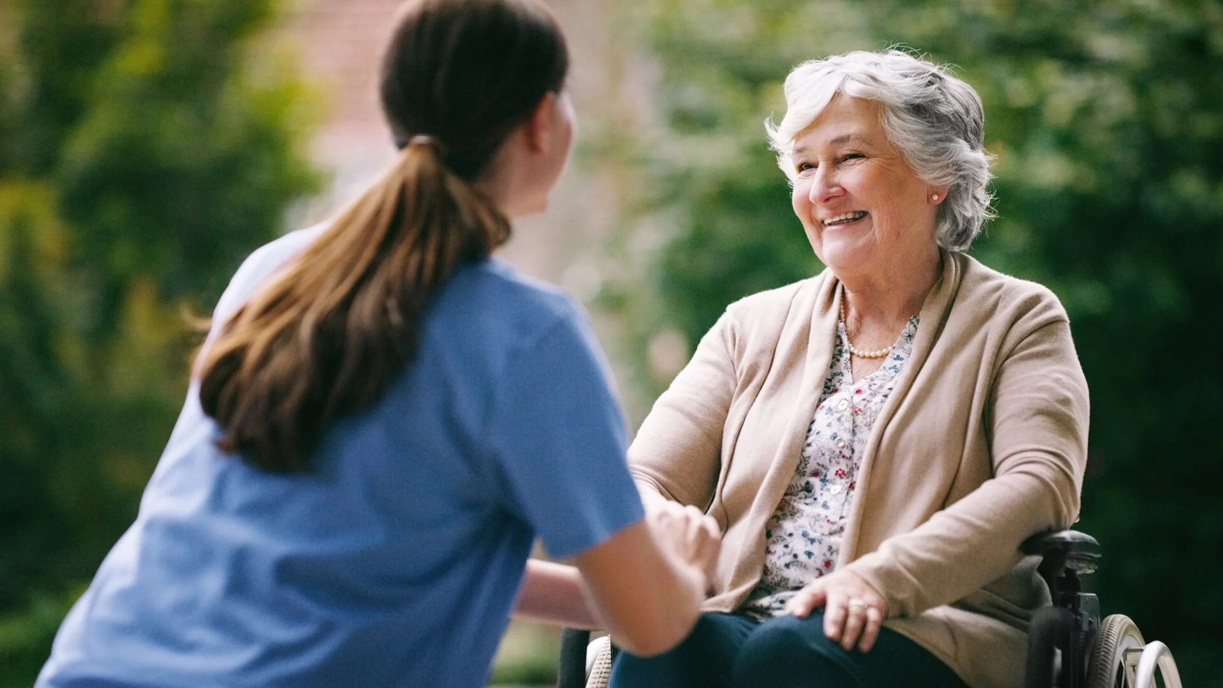 Elderly woman in a wheelchair outside with her nurse who is sitting down and holding the woman's hand.